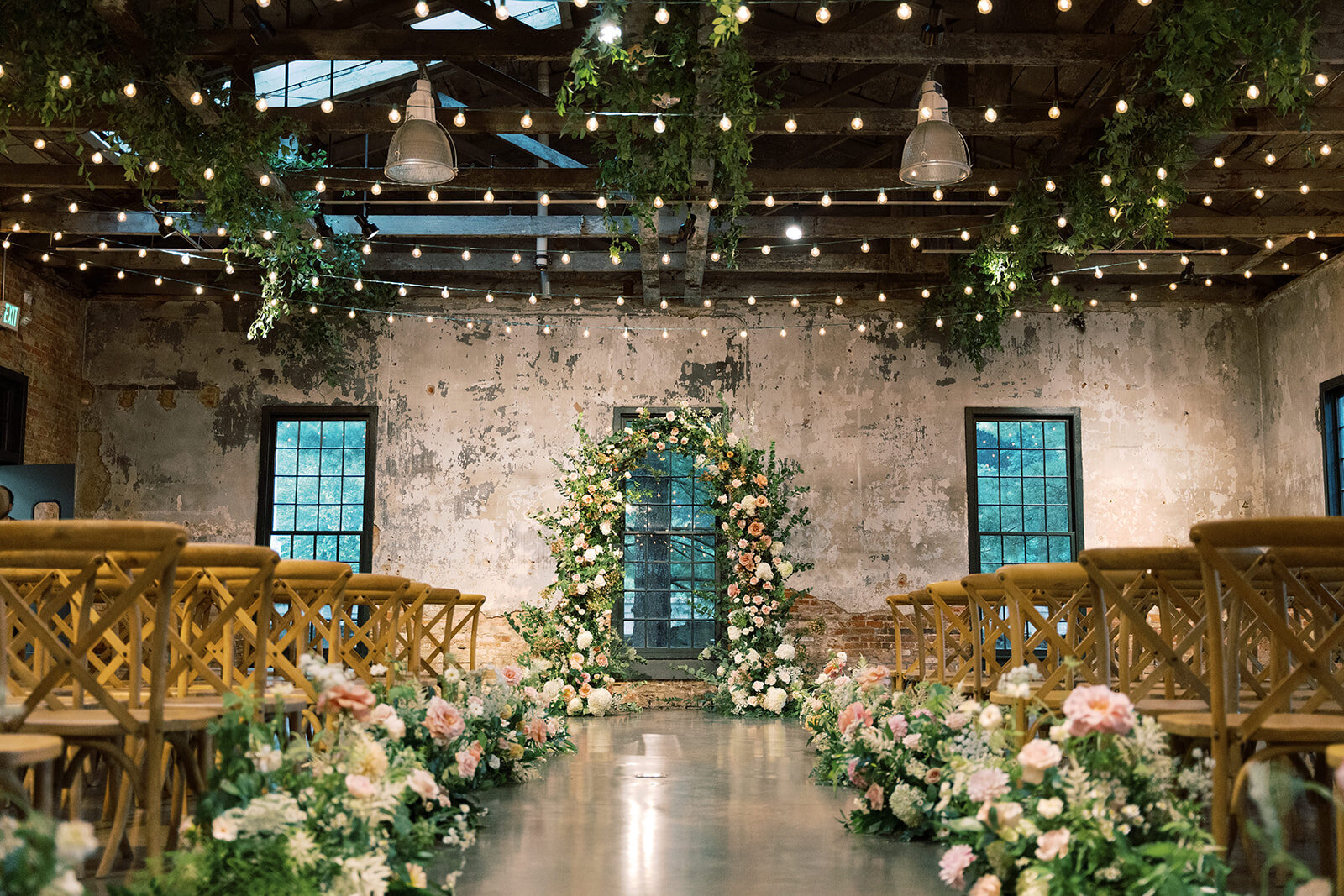 The ceremony inside the Washington mill dye house with a lush full floral arch and a meadow of florals going down the sides of the aisle including white, blush, and peach flowers and greenery hanging from the ceiling.