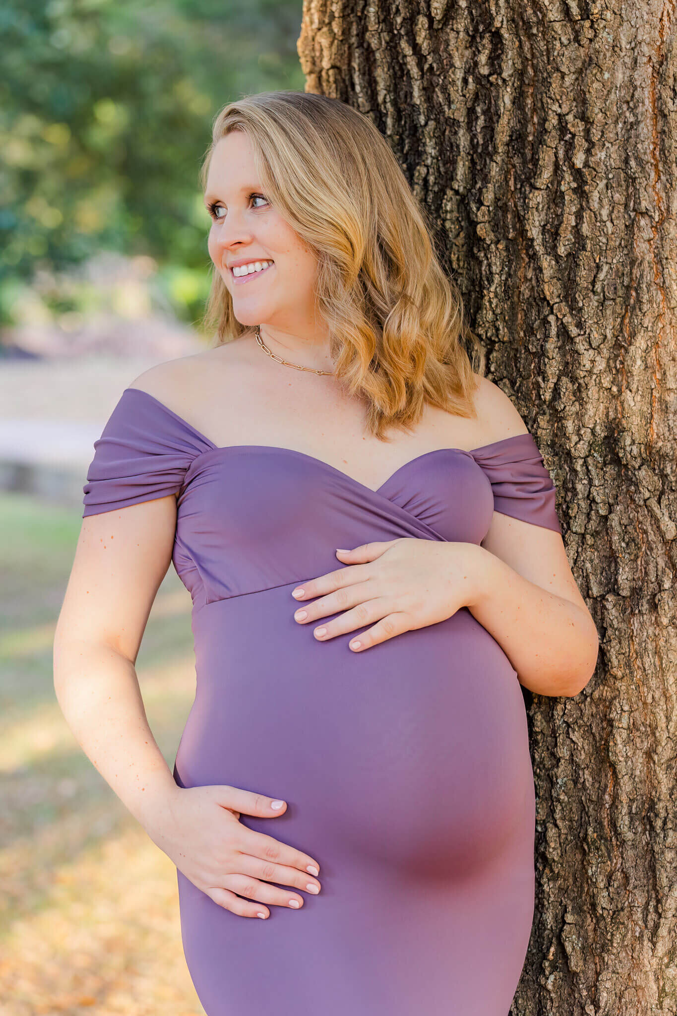 A maternity portrait of a woman in purple leaning against a tree.