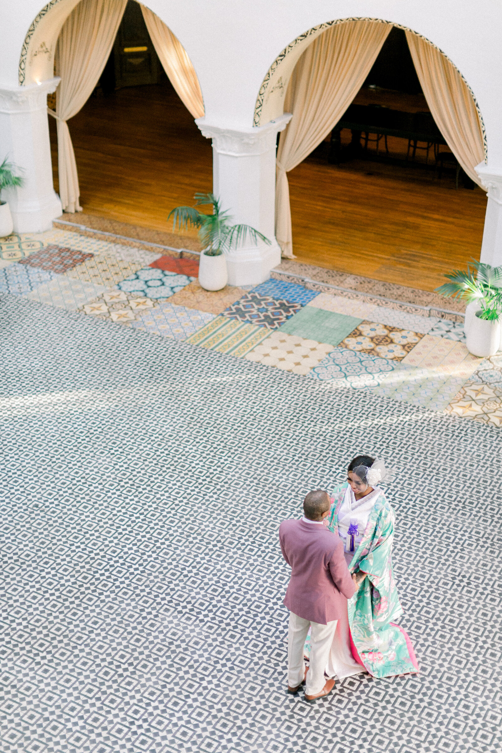 Bride in green floral wedding kimono with groom in mauve suit sharing a quiet moment beneath elegant Spanish-style arches at The Ebell Club Long Beach, kimono rental wedding photography | ロングビーチ・エベルクラブのスパニッシュ調アーチ下で寄り添う花嫁と新郎　グリーン色打掛の着物レンタル和婚撮影