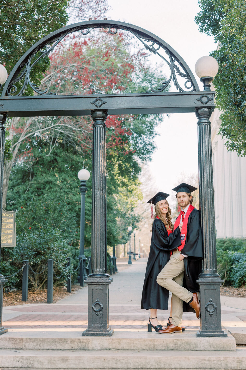 Girl and guy in cap and gown pose under UGA Arch