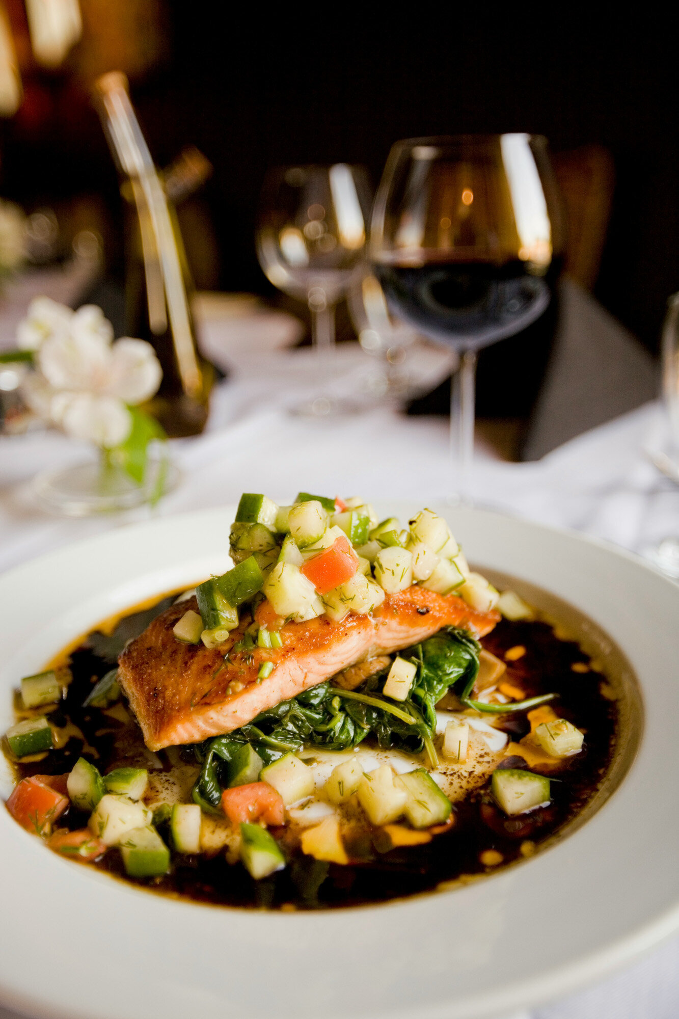 A gourmet dish on a table is elegantly set with wine glasses and a small floral centerpiece in the background.