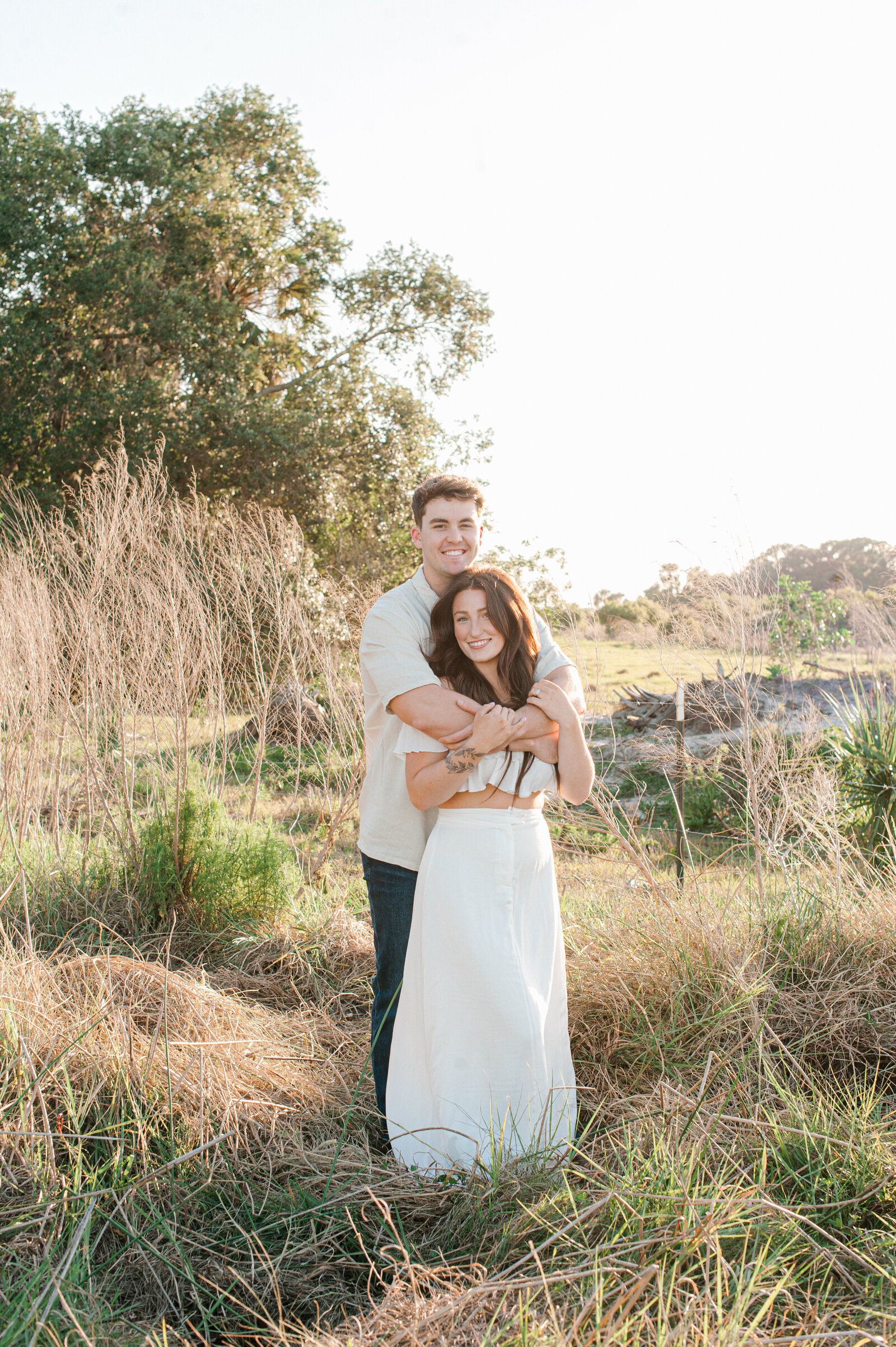 Couples stands in a tall grass field embracing during golden hour
