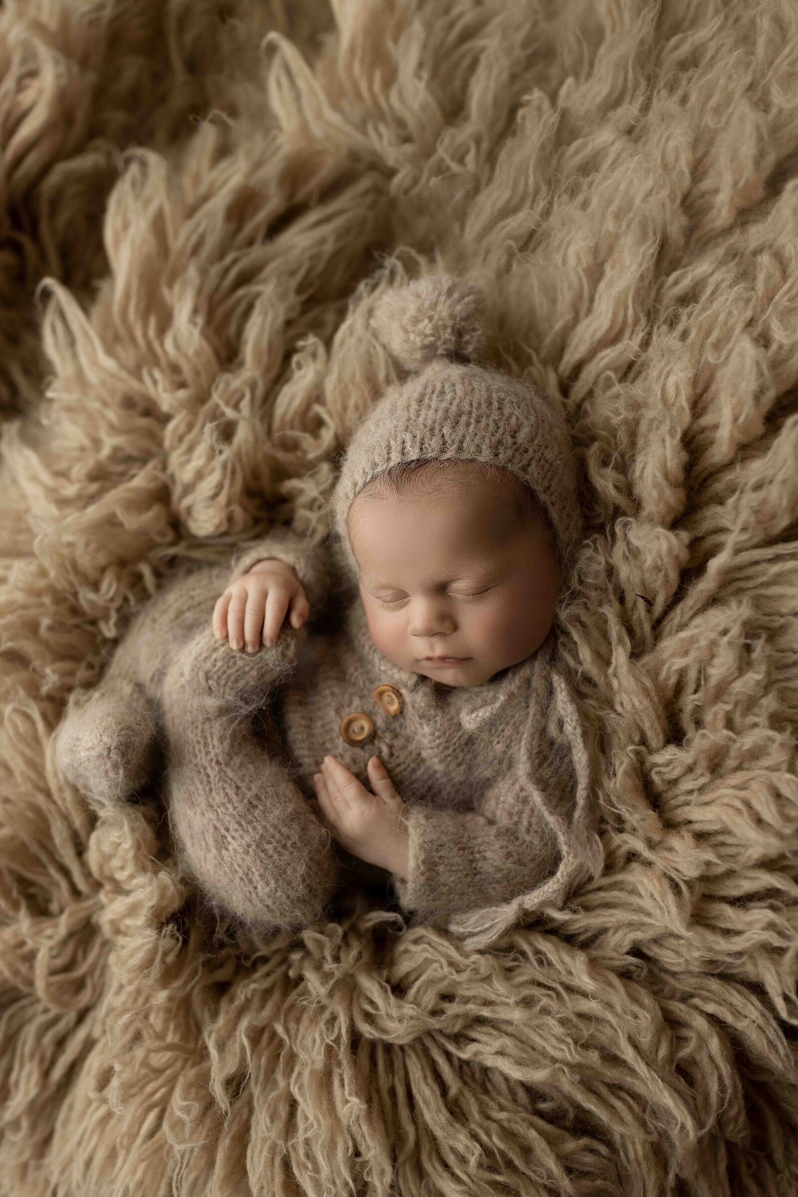 London, Ontario Best Posed Newborn Photography - Baby sleeping curled up on beige fluffy flokati wearing matching knit onesie and bonnet