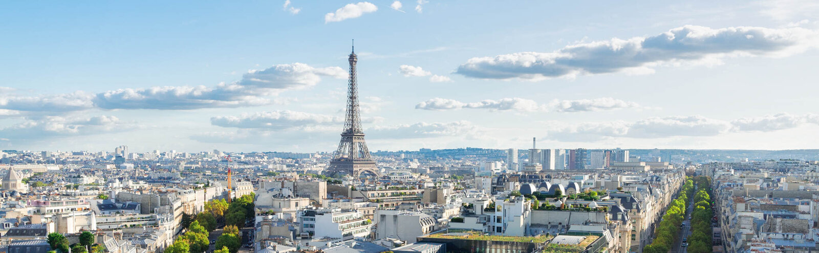 A panoramic view of Paris featuring the Eiffel Tower under a blue sky with scattered clouds. The cityscape includes various buildings, greenery, and wide streets.