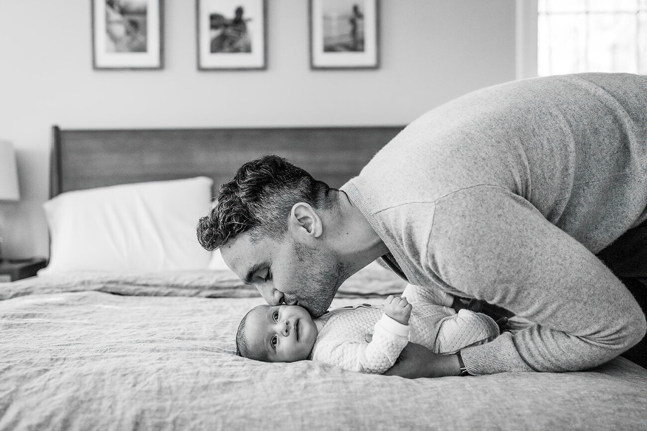 black and white photo of new father bending over to kiss newborn laying on bed