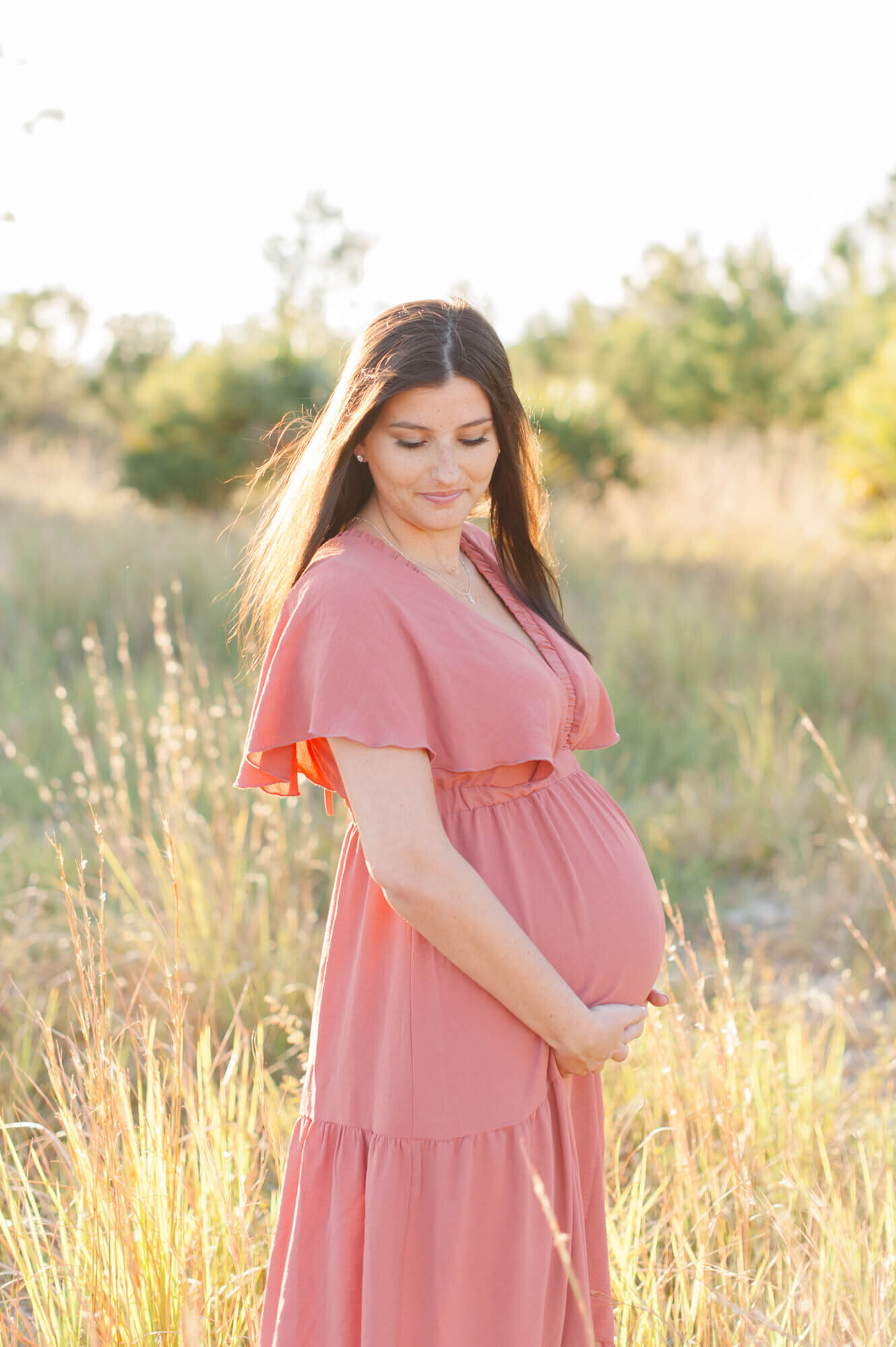 New mom holding her belly in a tall grass field at sunset wearing a pink dress