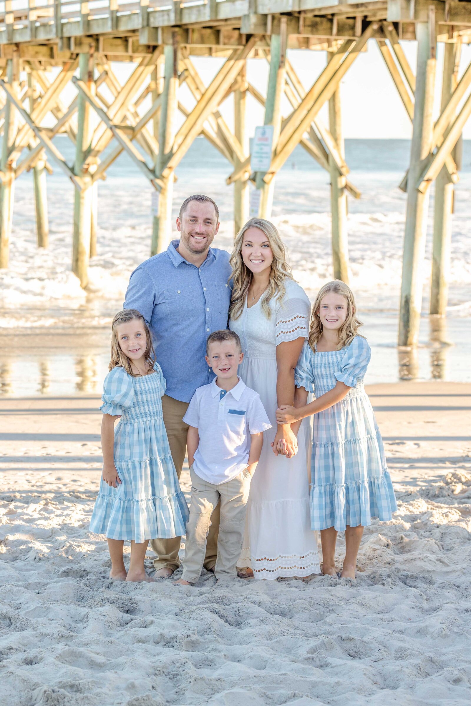 A family of five stands under a pier, dressed in blue and white outfits. The ocean waves and sandy beach provide a picturesque backdrop. Ideal for destination family photographer NC, showcasing elegant and serene beachside portraits.