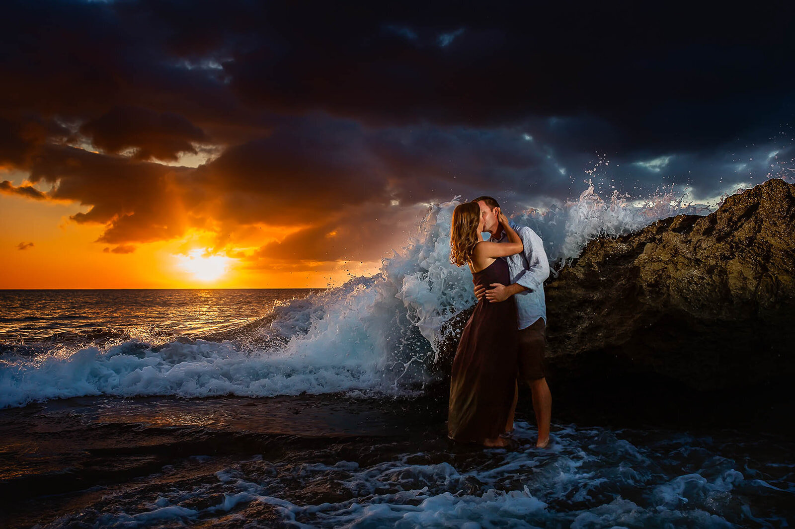 A couple sharing a kiss on a rocky shore as the sun sets behind them, captured by San Antonio luxury wedding photographer CMH Studios
