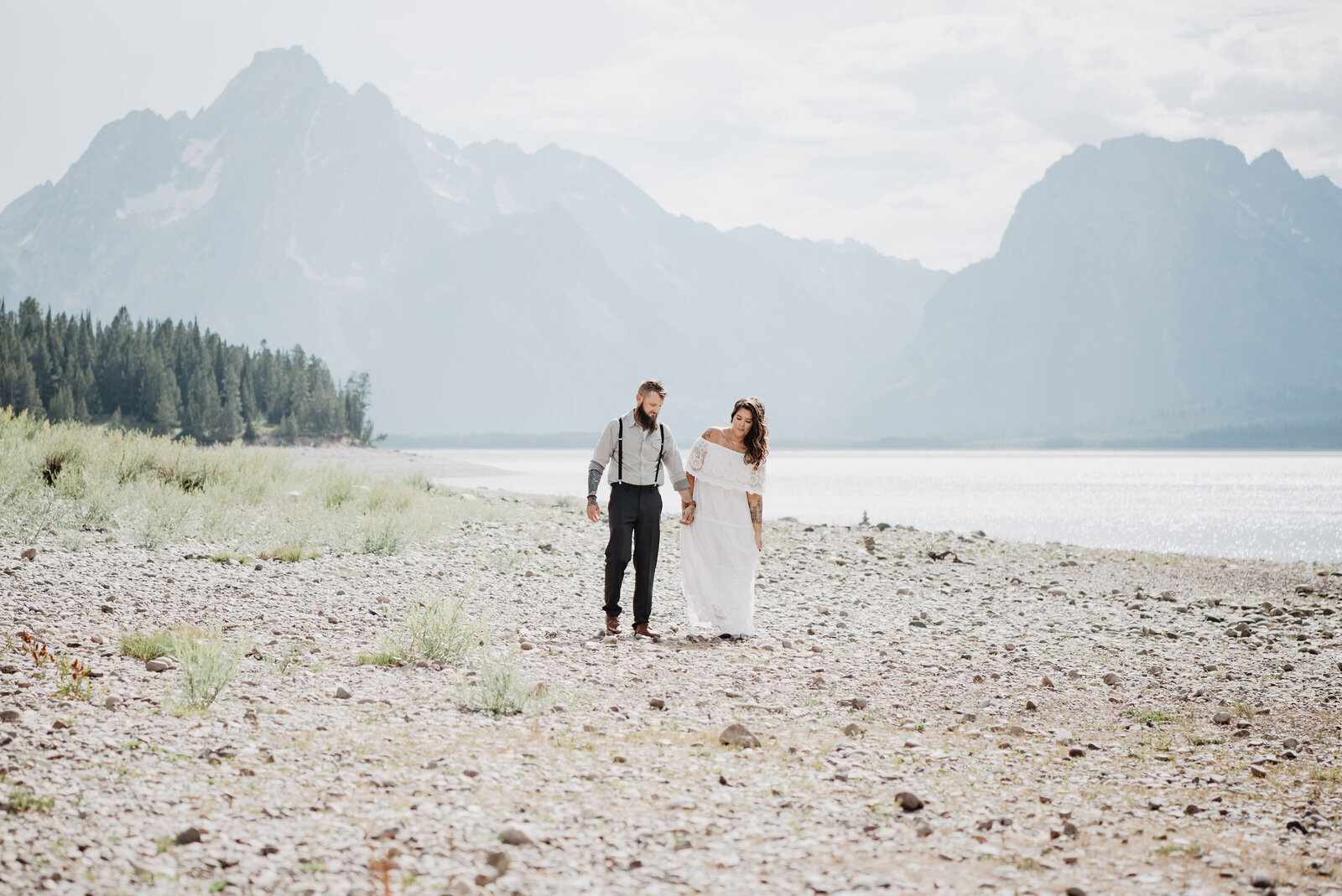 outdoor jackson hole wedding with bride and groom holding hands and walking on the shore of the beach in the grand tetons with the mountains towering over water