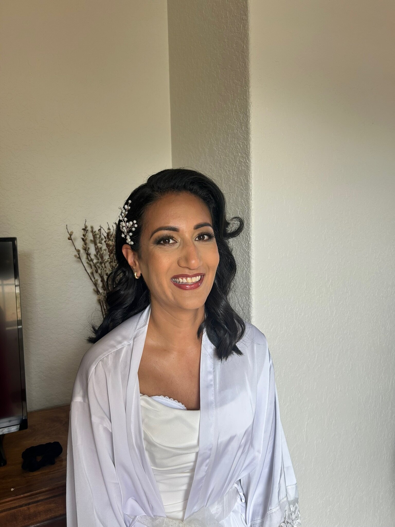 The woman in a white robe, with styled dark hair adorned with elegant accessories, exudes joy indoors. It's a glimpse of the bridal beauty experience Texas offers, where every detail is designed to make her smile as radiant as her beauty transformation.