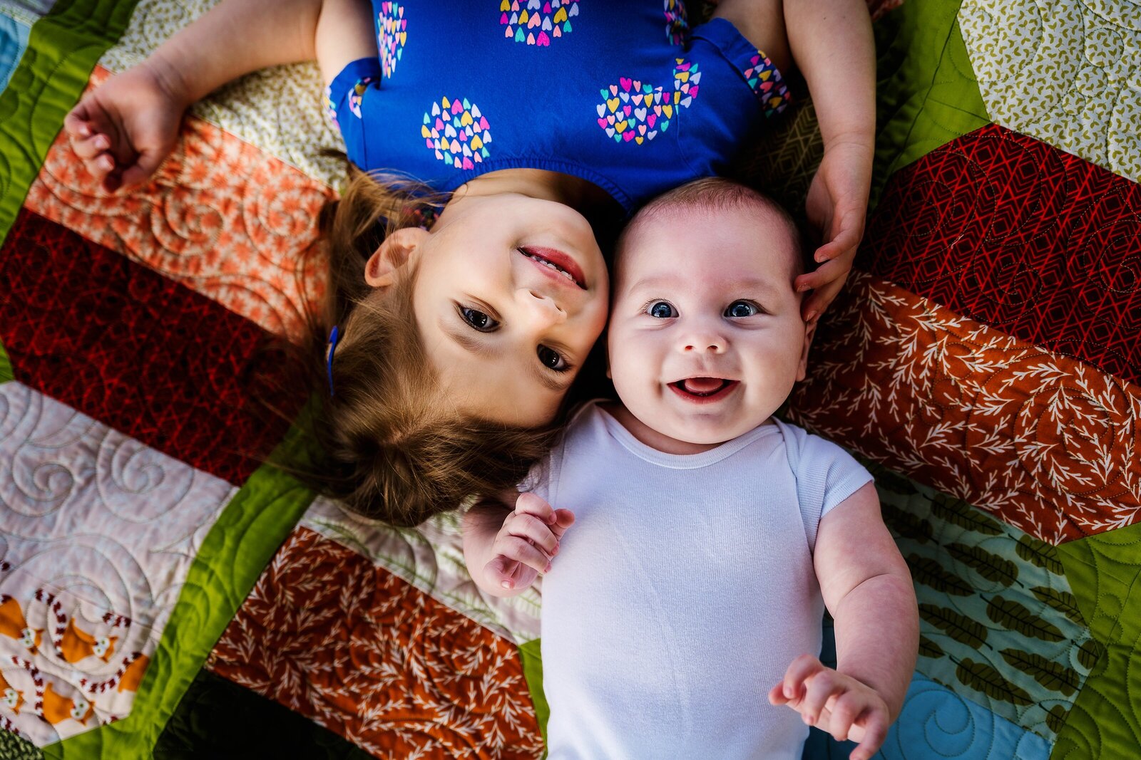 Toddler and baby smiling at camera on colorful quilt