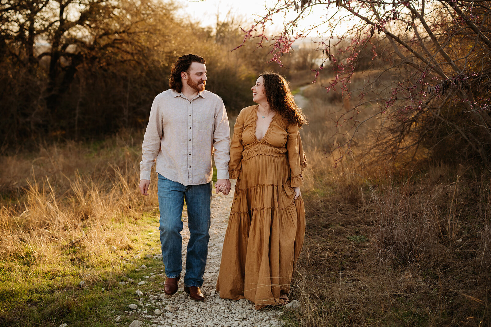 Husband and wife walking down trail at Lake Worth while Paula Goforth captures their spring maternity session. Mom is wearing a long mustard dress by reclamation and dad is wearing jeans and a button down shirt. 