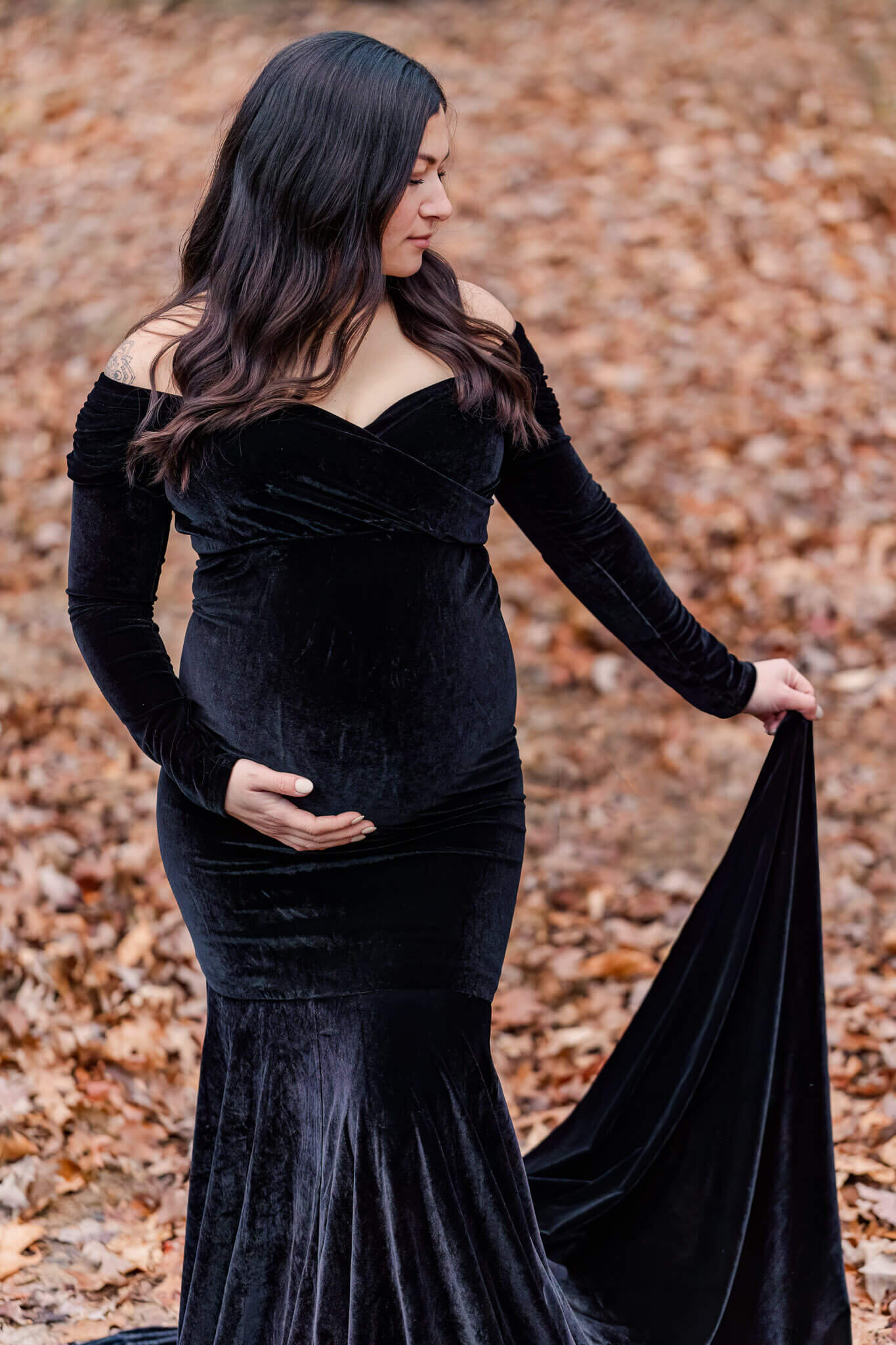 A mother-to-be posing in a black dress among the leaves at a park in the fall.