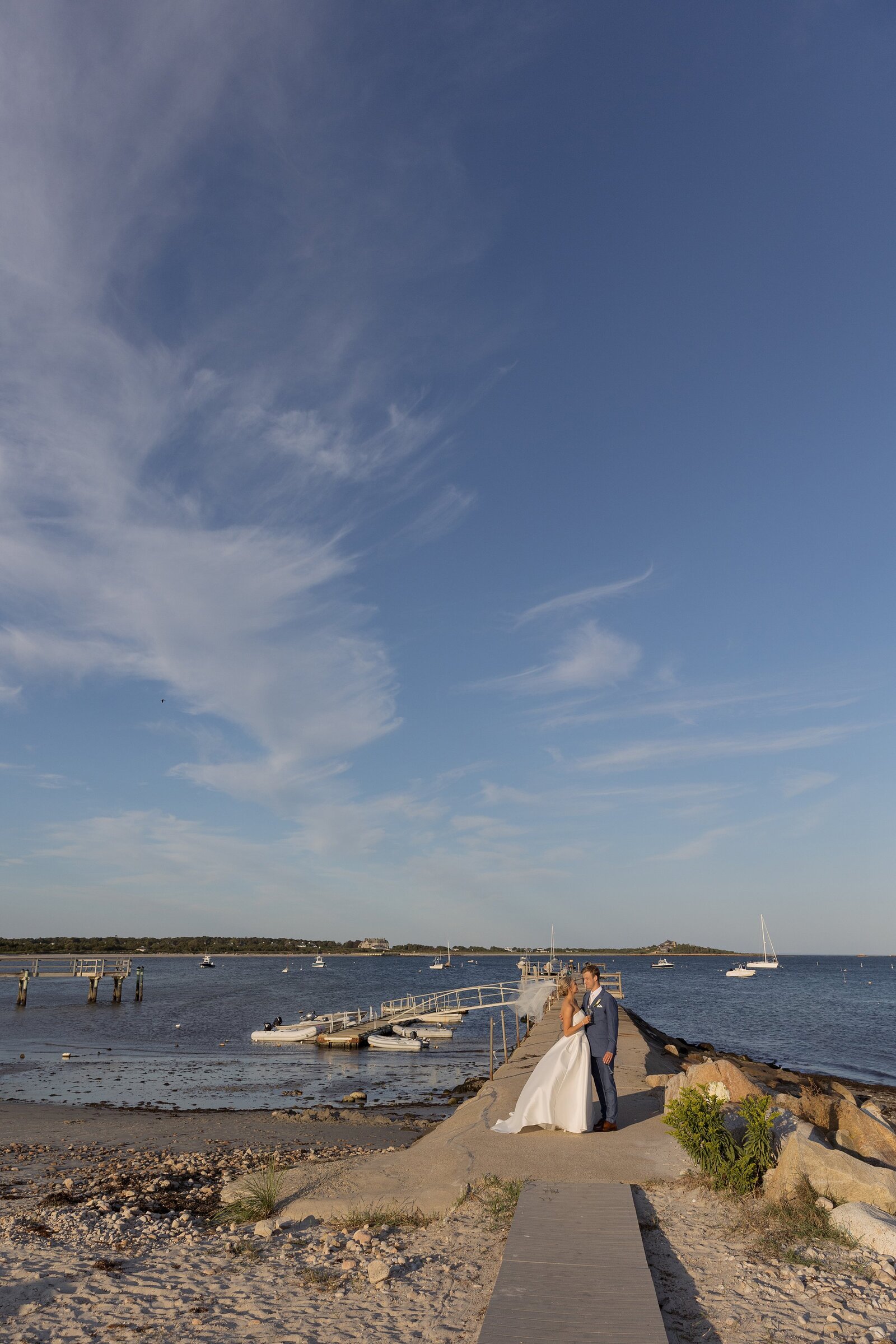 Bride and groom stand on rock jetty at sunset. 