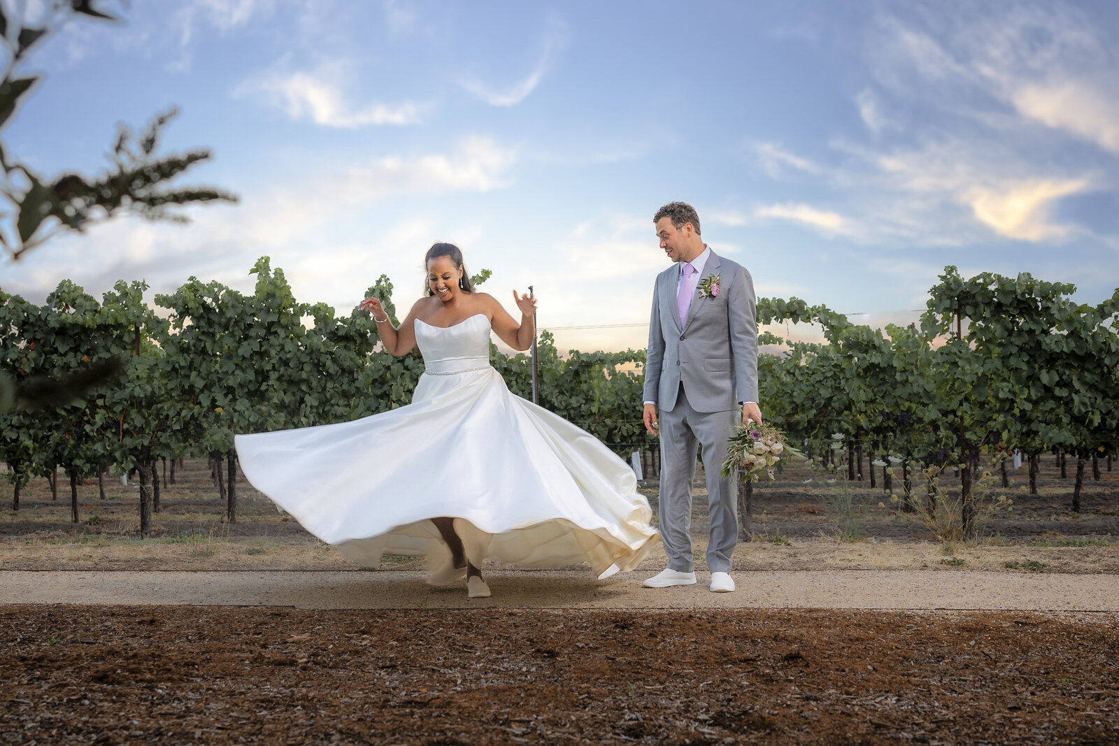 Sacramento wedding photographer capture bride twirling her dress in front of a vineyard and groom watches.