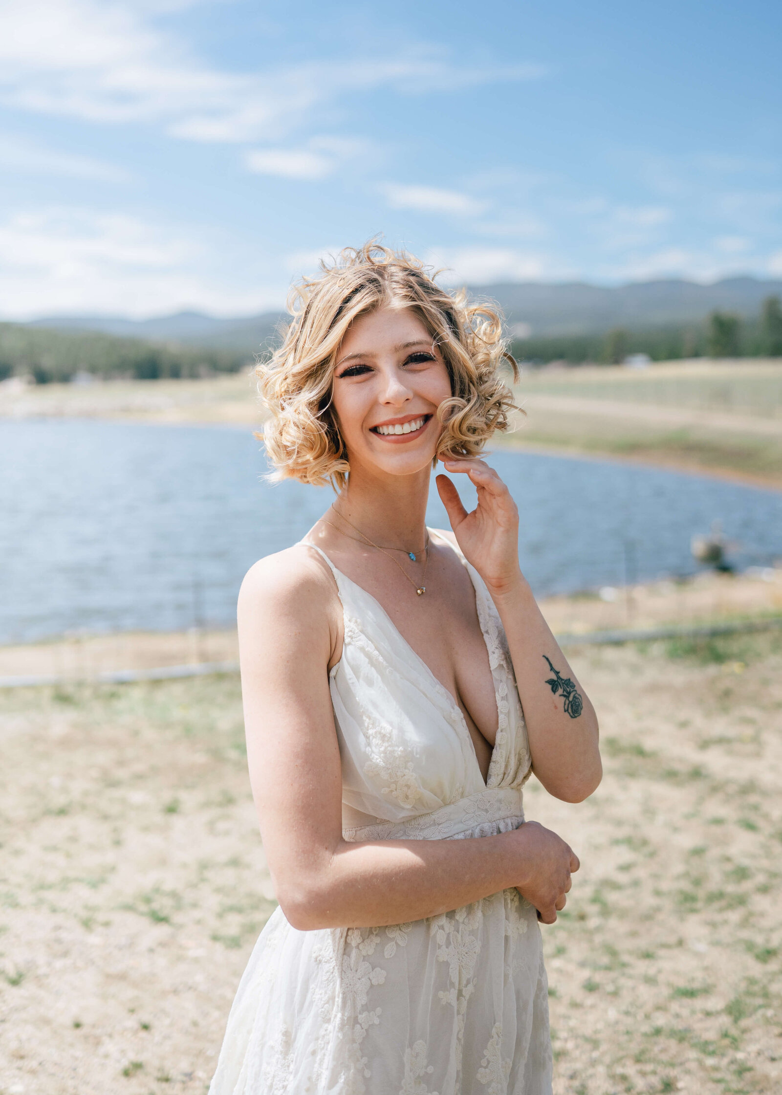 Blonde bride with short curly hair stands in front of a pond and smiles at the camera during her bridal portraits