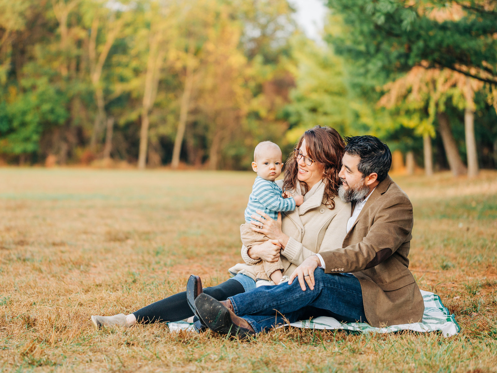 Young family sits on a blanket in a fall clearing looking at their young baby