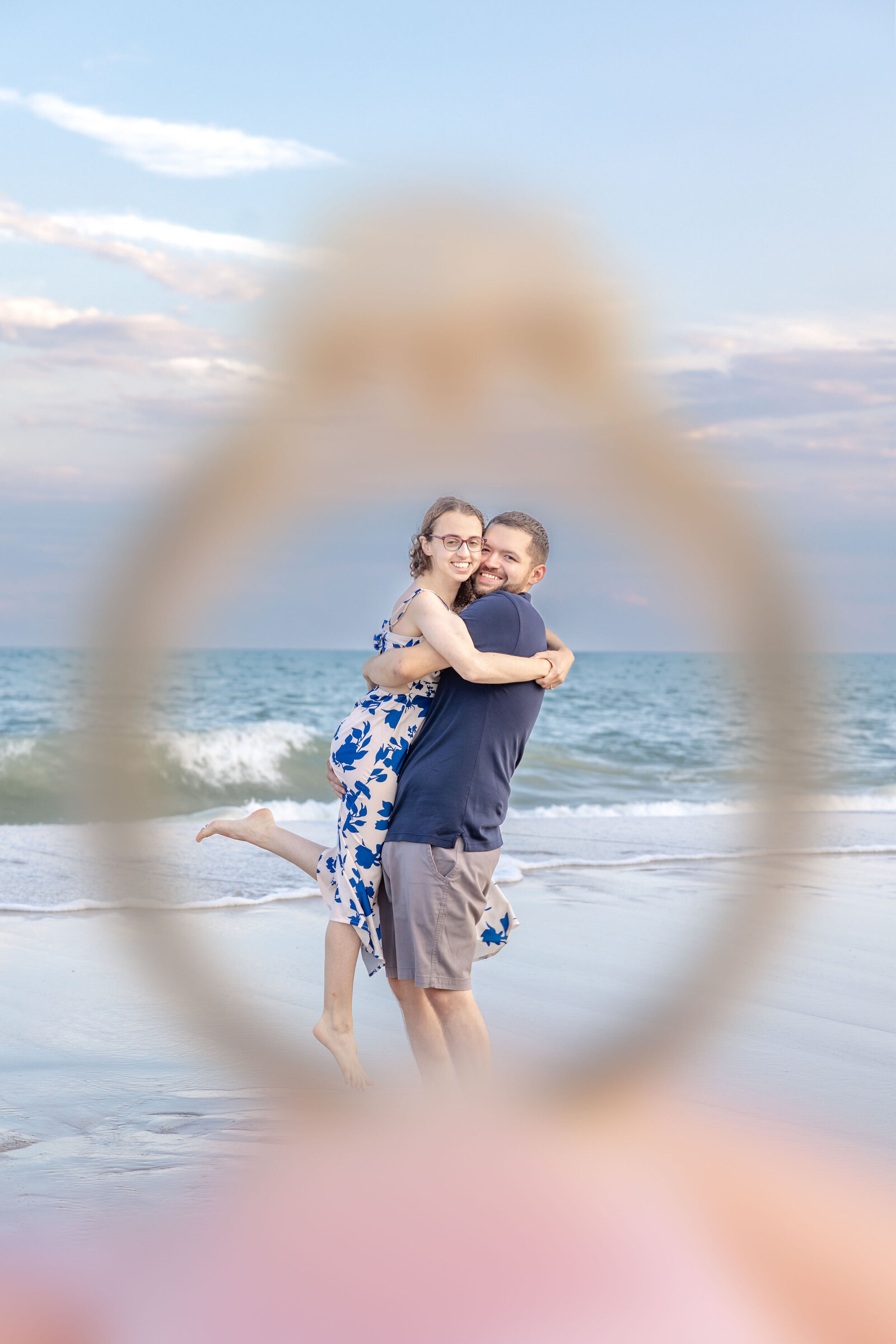 A joyful couple on the beach in North Topsail Beach, NC, framed creatively through an engagement ring. This image captures the essence of unique wedding photography, ideal for couples seeking artistic wedding photography and videography services in North Carolina.