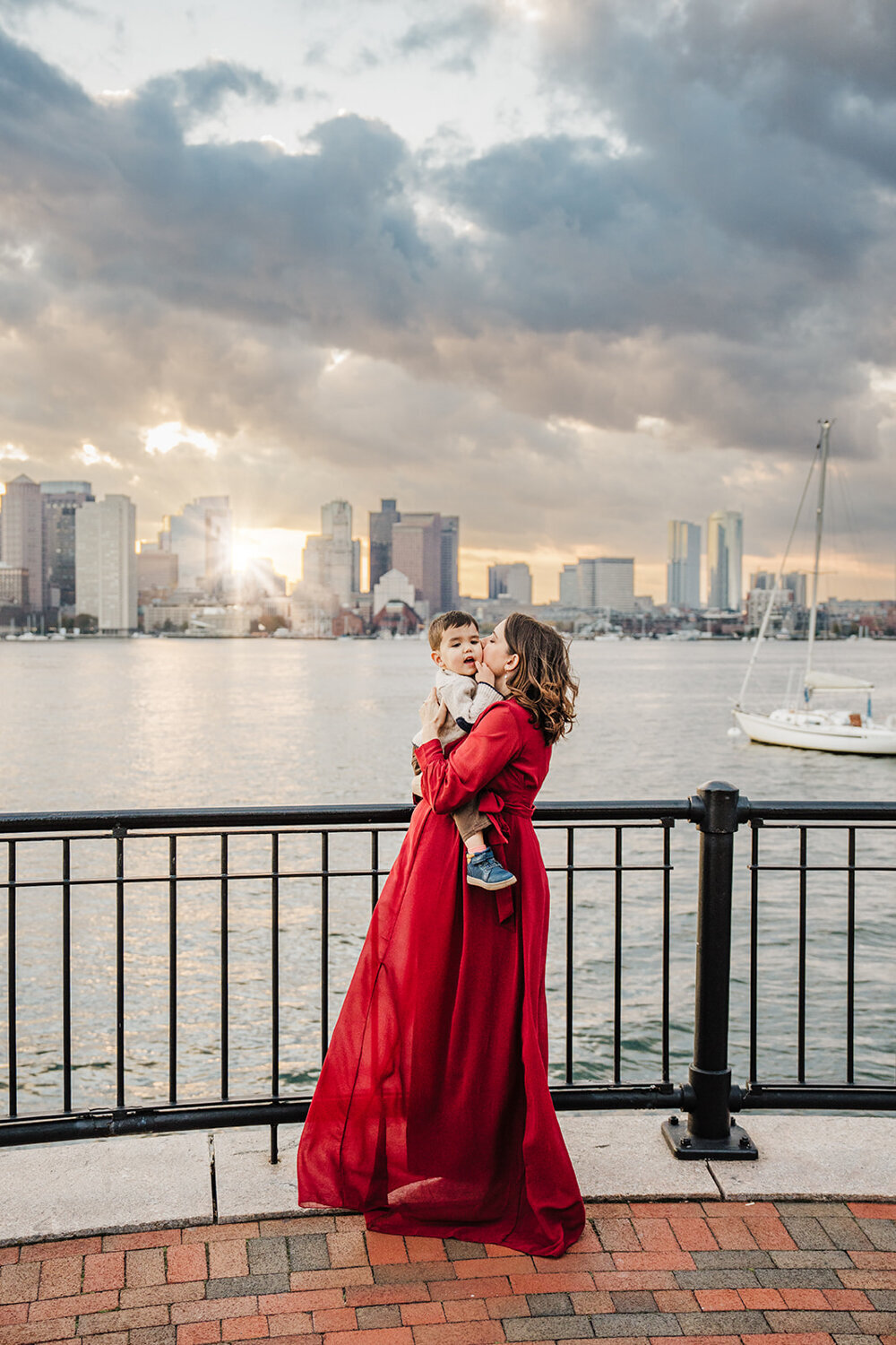 pregnant mom in red dress kisses toddler son with sunset city skyline backdrop
