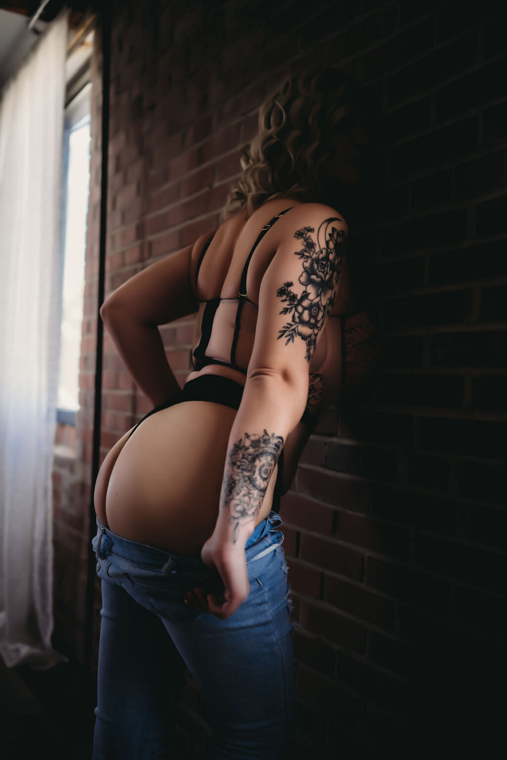 woman with tattoo on arm in black bra and jeans leaning against brick wall showing butt