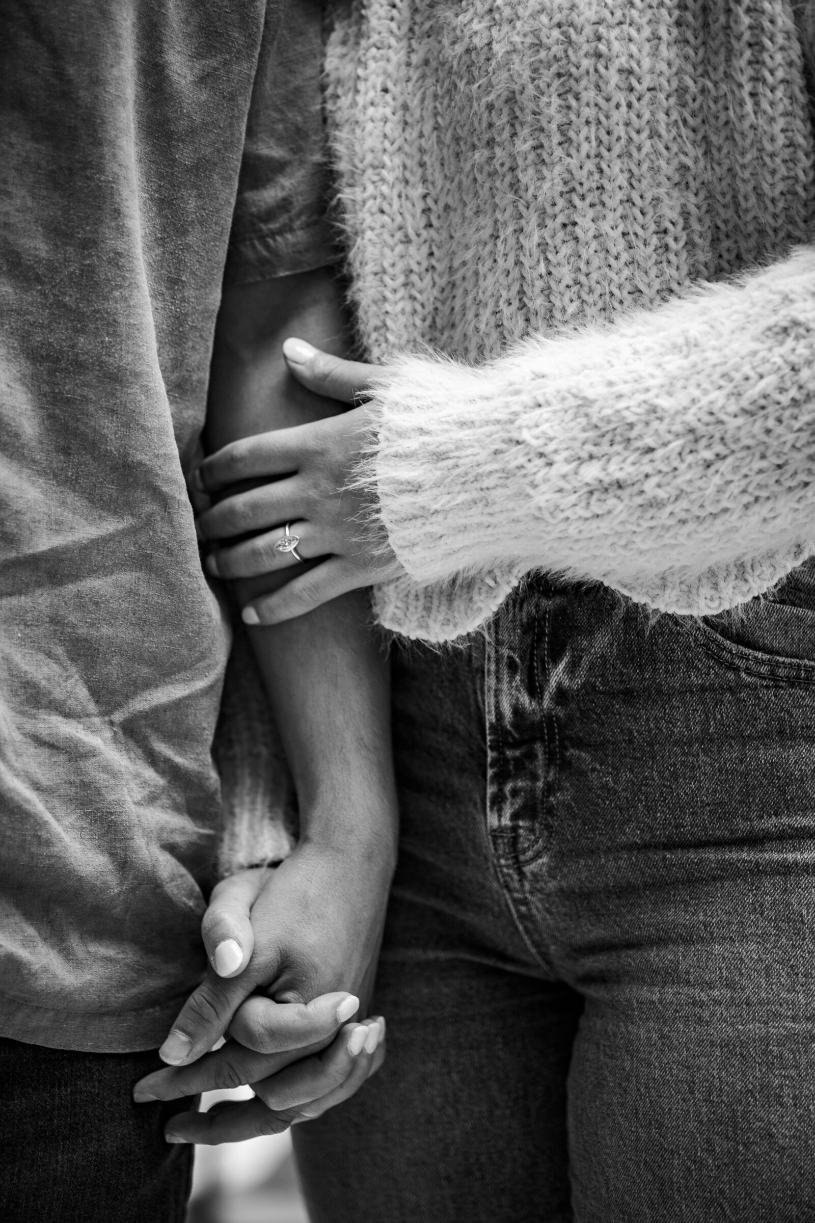 Black and white shot of a man and woman holding hands during their engagement session, with focus on the woman's engagement ring.