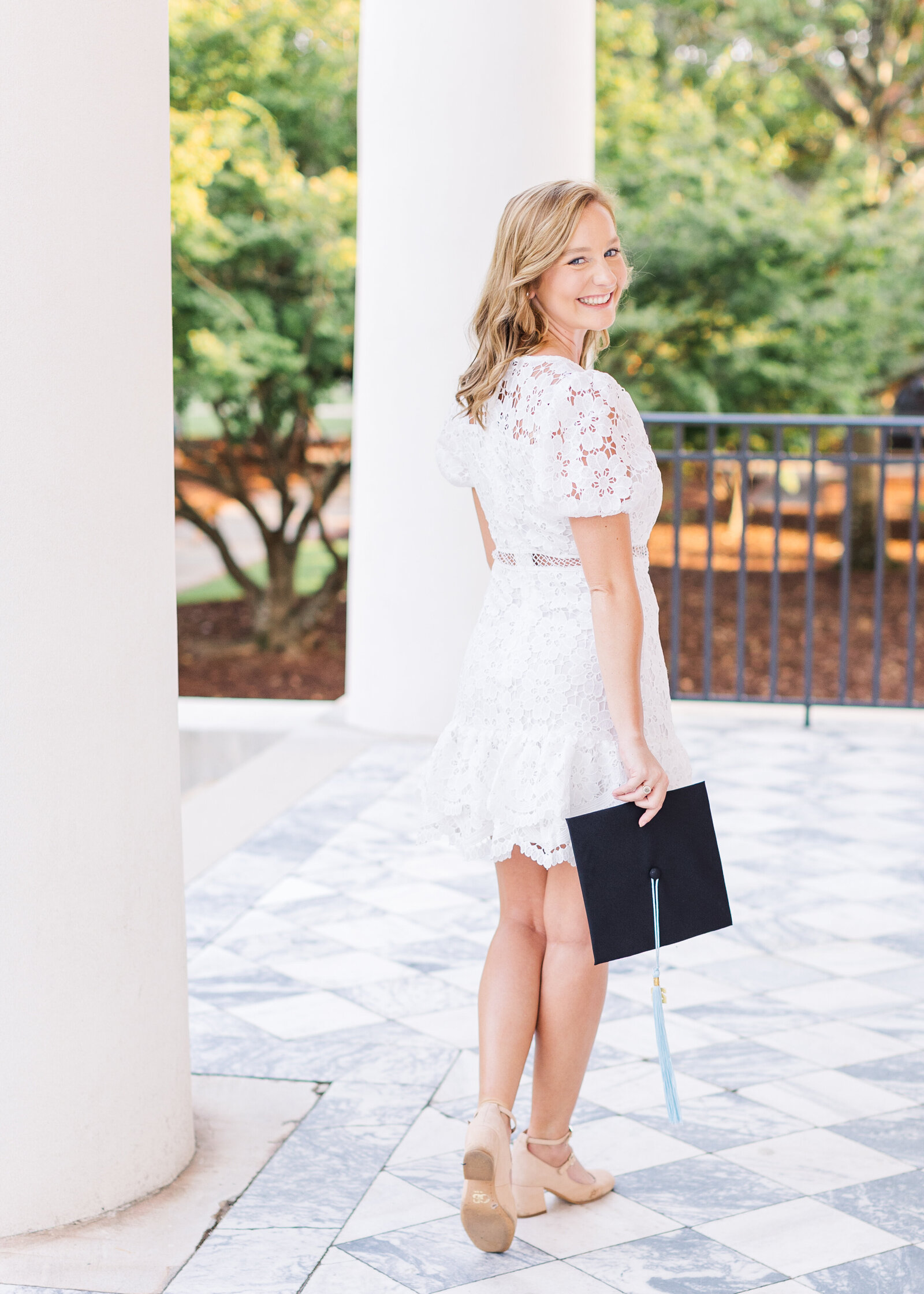 Shelby smiling while holding her graduation cap, wearing a white lace dress, at a classic columned building during her senior portraits by Abby Bell Photography.
