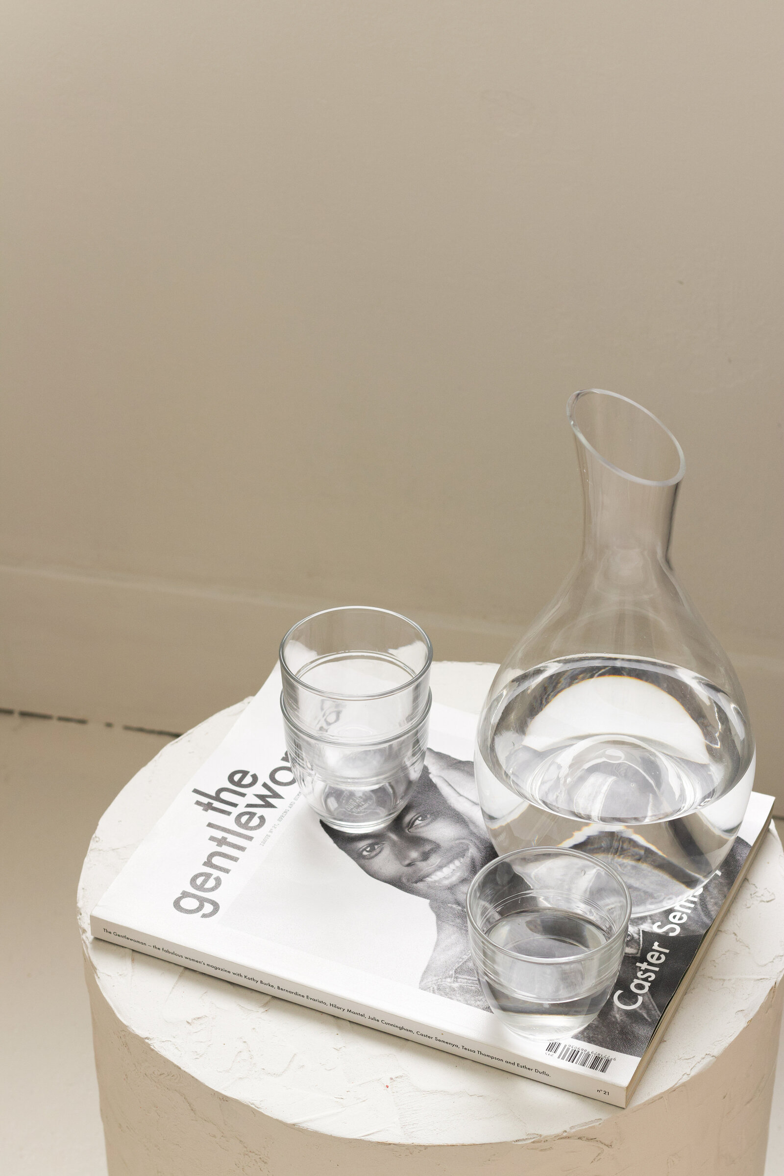 A glass water bottle and a transparent glass on a magazine. Beige background
