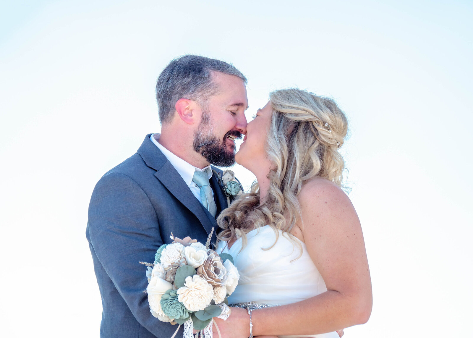 Groom leans in for kiss as bride giggles in Wilmington, NC. OR  Bride and groom sharing a sweet kiss at their beach wedding, with a clear sky as the backdrop, captured in North Carolina. 