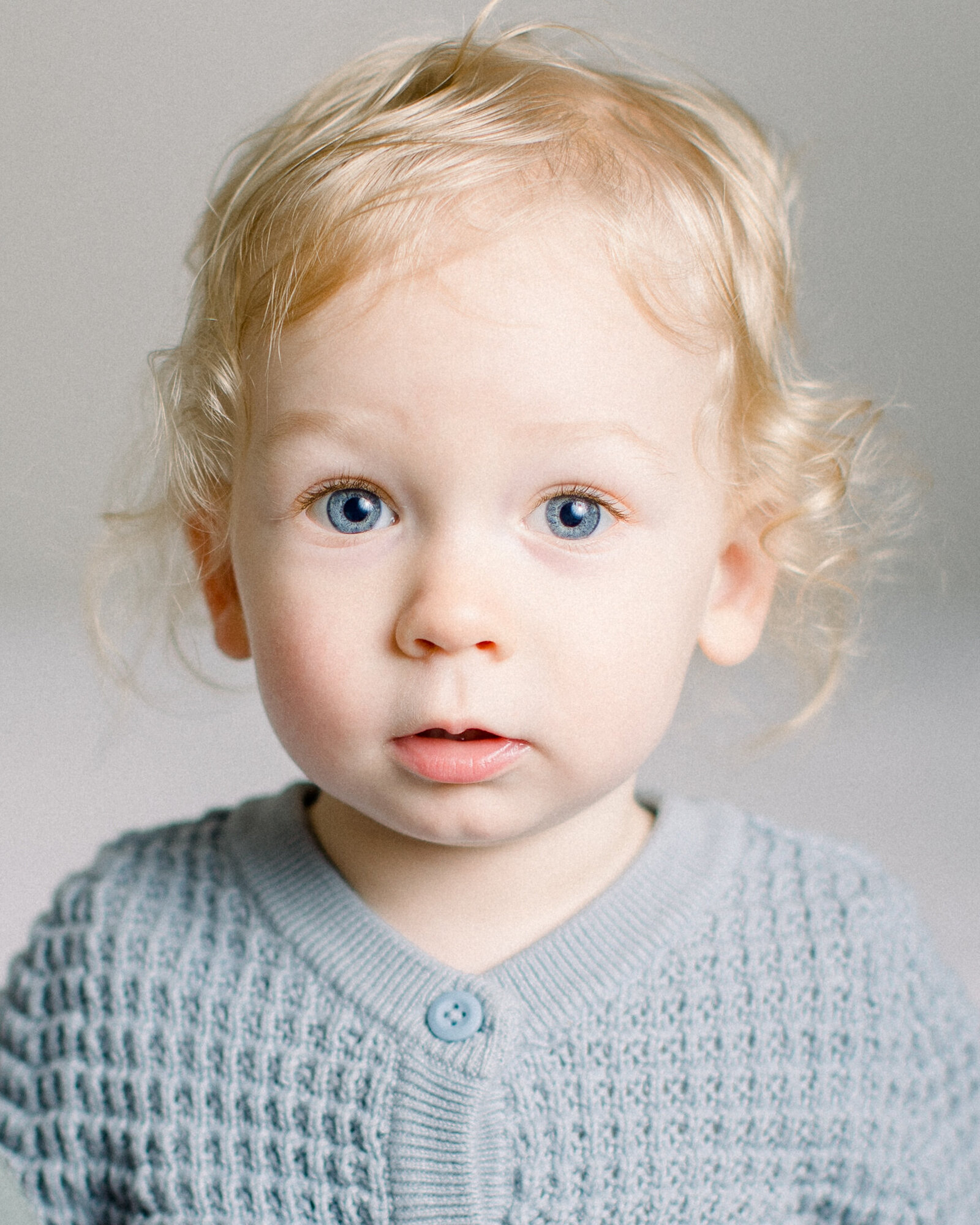 One year old boy in blue cardigan with blonde curls and blue eyes looking inquisitively at the camera by NH newborn photographer Fieldstone Studio.