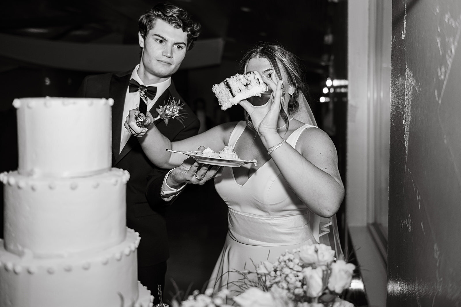A fun and candid moment as the bride playfully holds up a slice of cake while the groom watches during their wedding reception, captured in black and white by CMH Studios