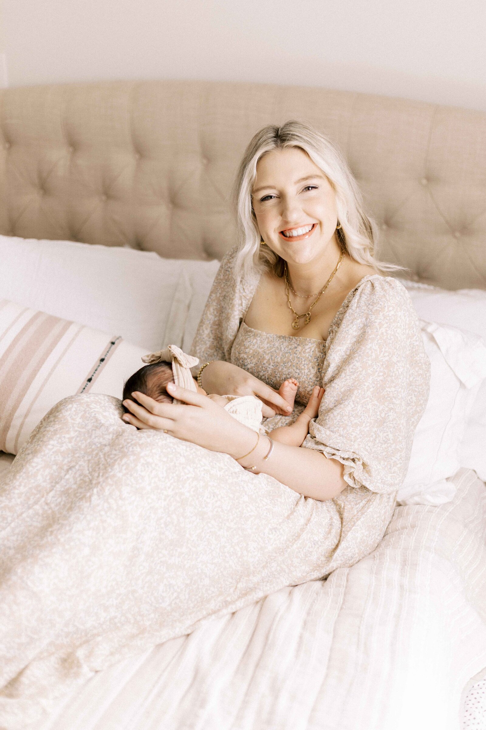 mom sitting on bed with baby on her knees during newborn photos in peoria illinois 