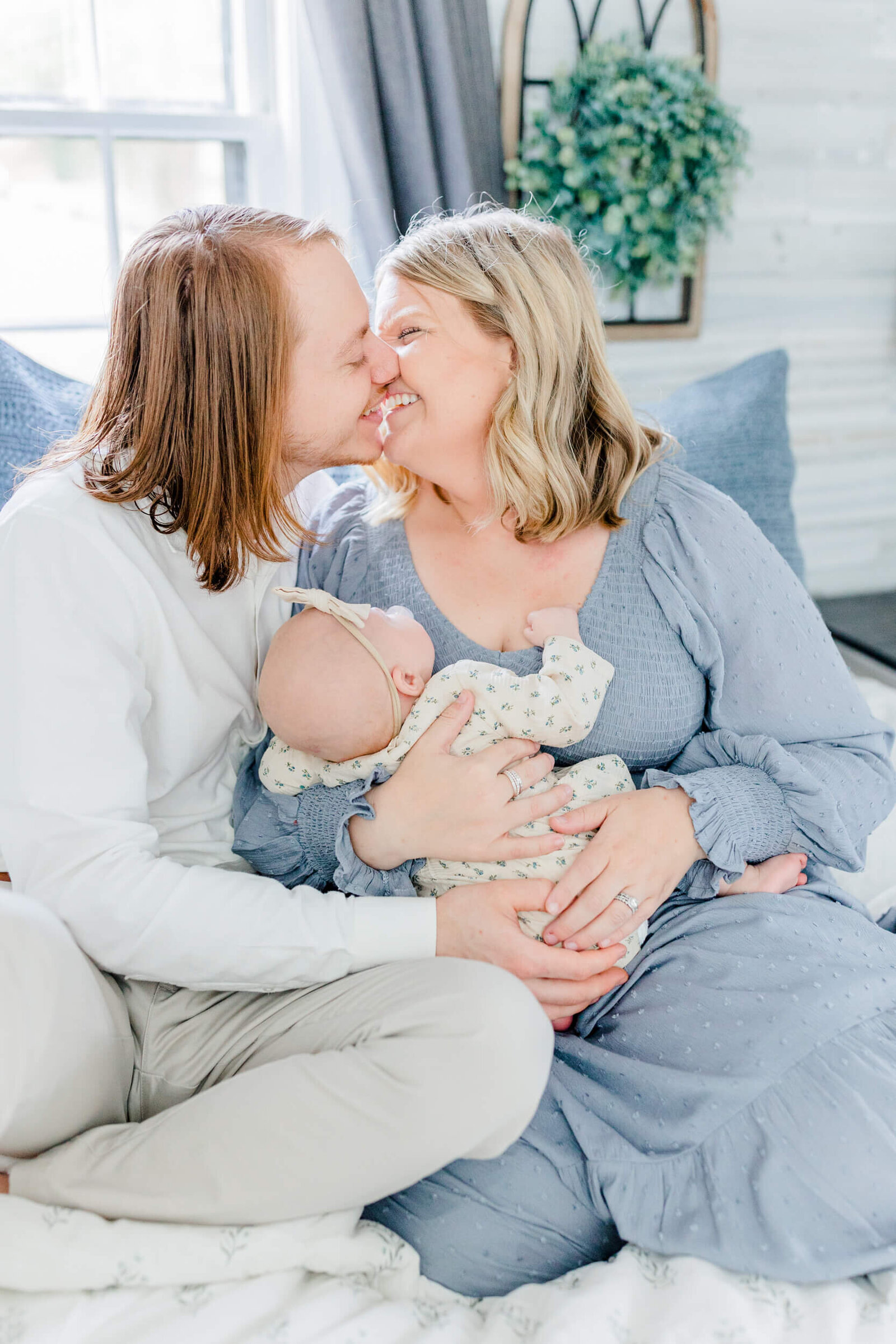 Mom and dad smile and lean in for a kiss on the bed while mom holds newborn