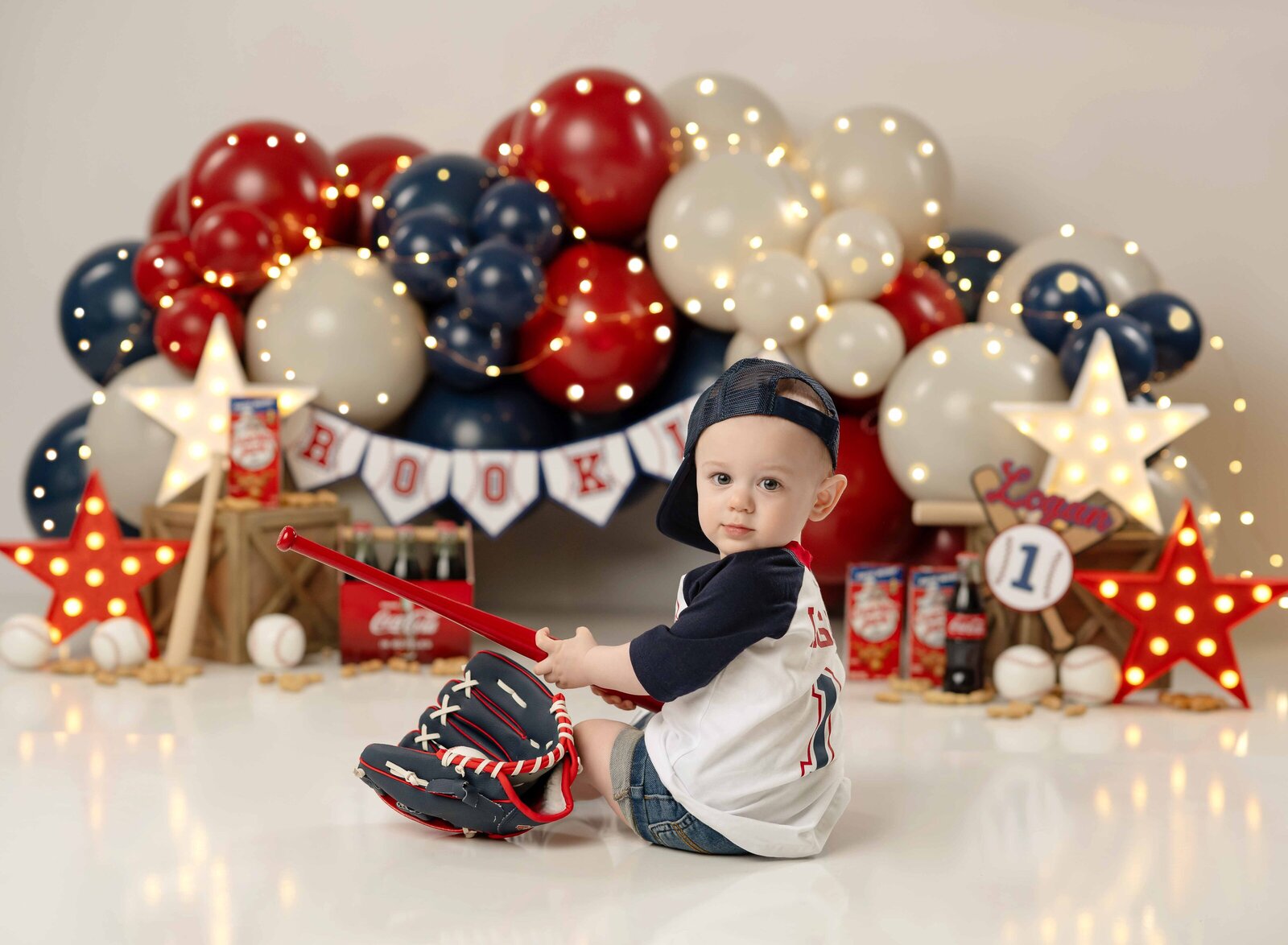 Baby on a baseball themed birthday set with a red, white and blue balloon garland. Baby is holding a baseball bat and looking over his shoulder.