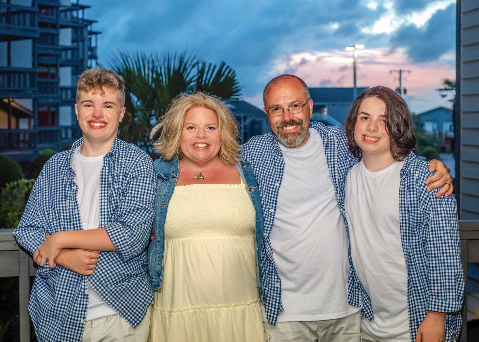 A family of four poses together at sunset, with beach homes in the background. Dressed in matching blue and white outfits, they capture a warm and intimate moment, ideal for destination family photographer NC, highlighting family unity and coastal living.