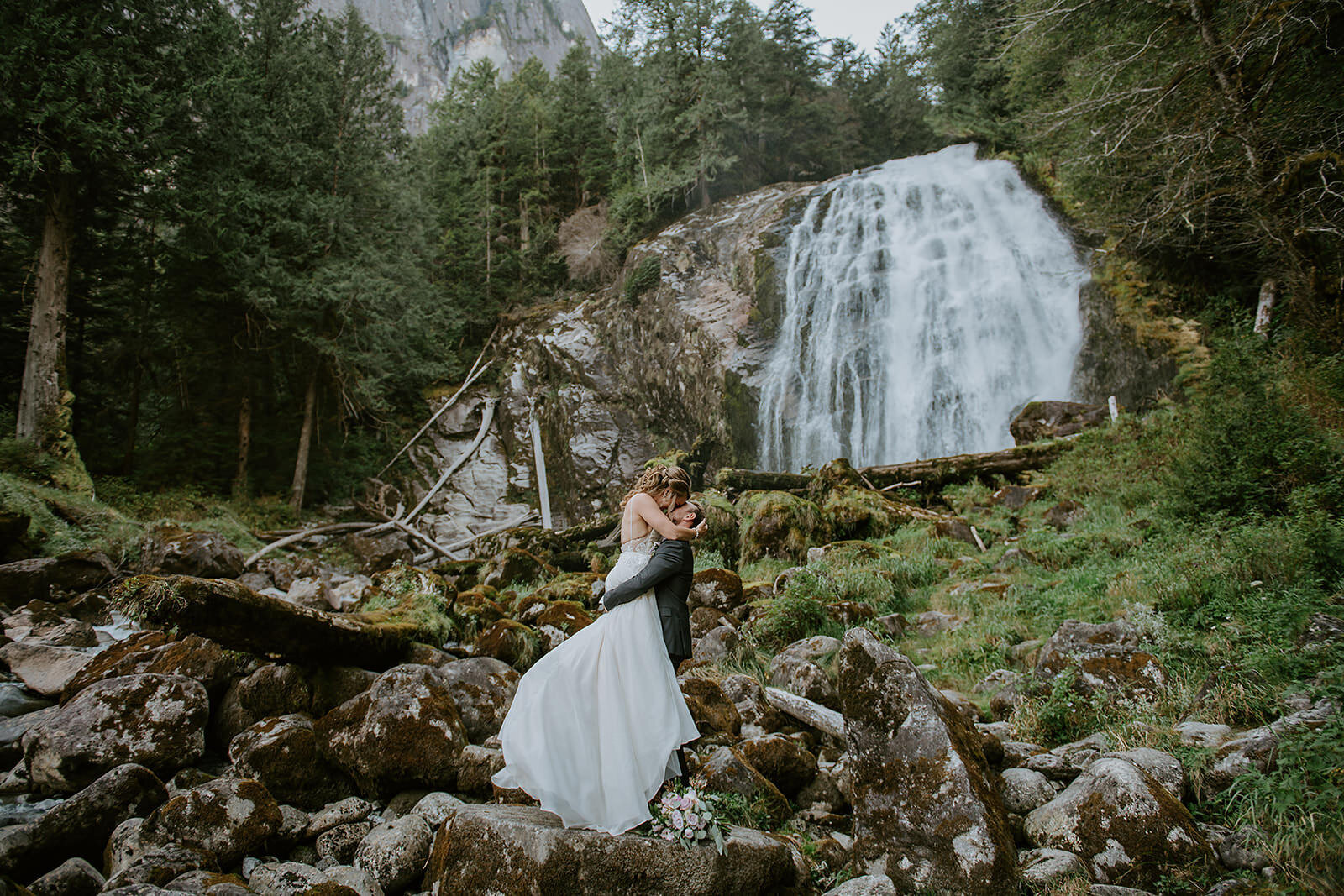 Bride and Groom in front beautiful Chatterbox Falls in British Columbia Canada at their romantic elopement. Couple sharing first kiss in the mist from the majestic waterfall.