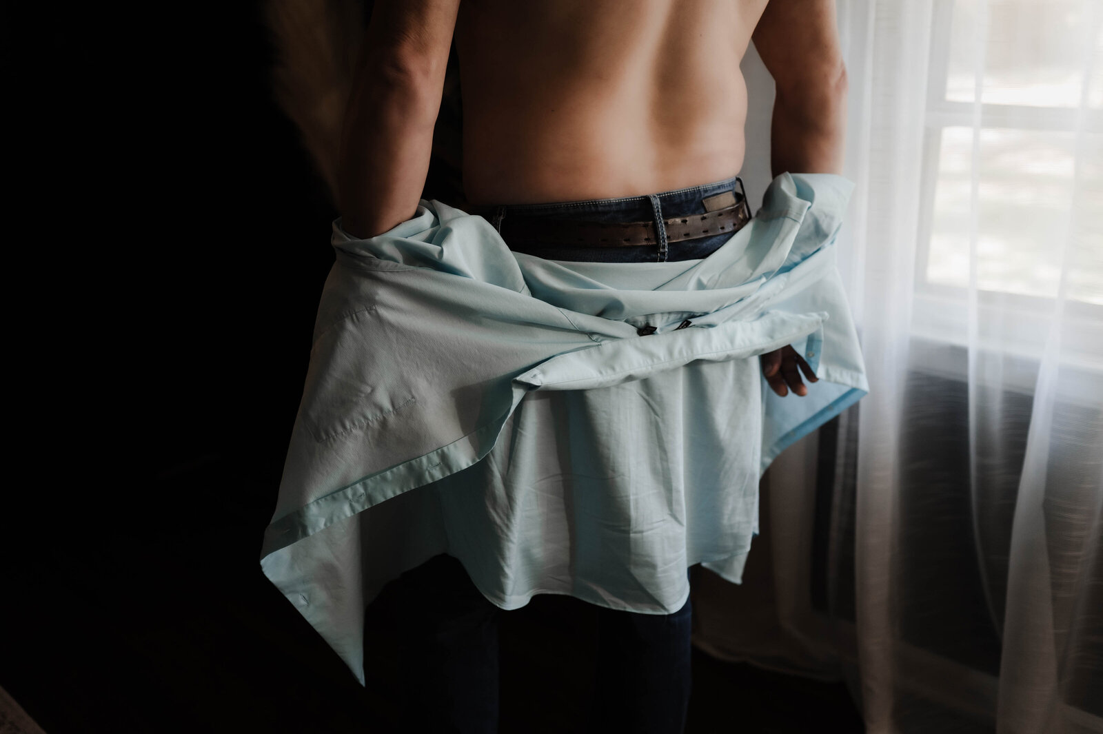 A man takes off his blue shirt while standing in a window