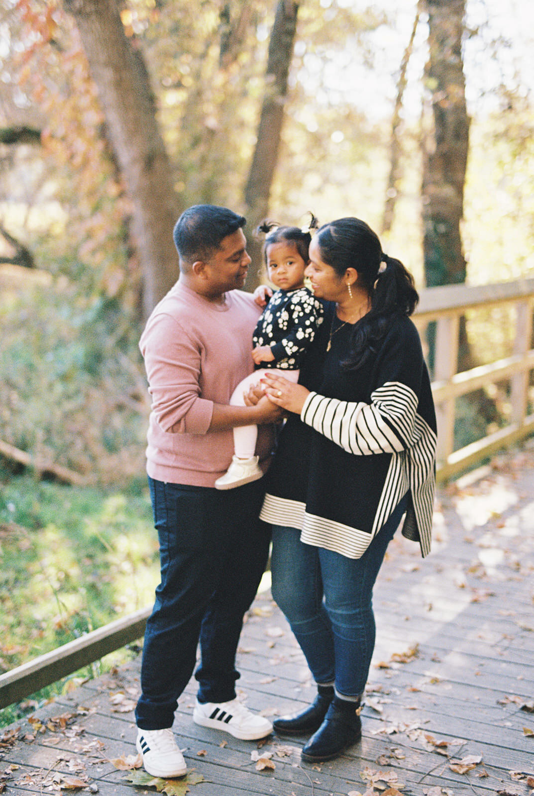 A sweet outdoor portrait of a couple holding their young daughter on a wooden path surrounded by autumn foliage.