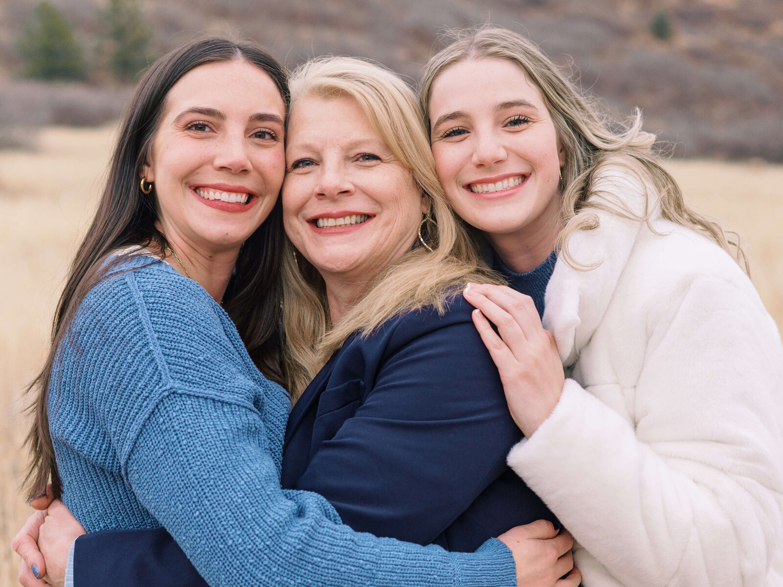 A blonde mother in a navy sweater receives a hug from her two adult daughters