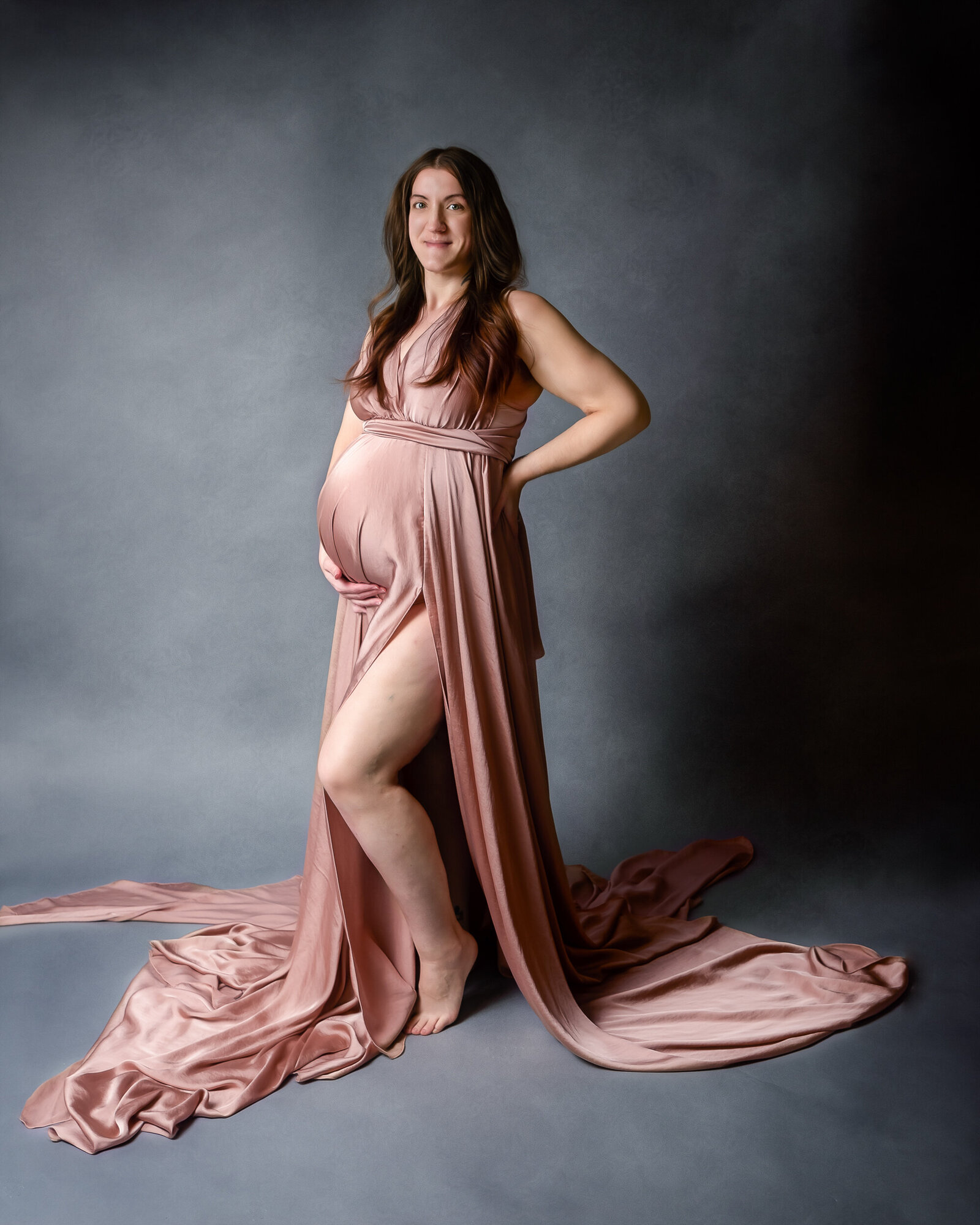 An expectant mother stands gracefully in a soft pink flowing maternity gown, her hand resting on her hip as she gazes confidently at the camera against a moody gray backdrop.