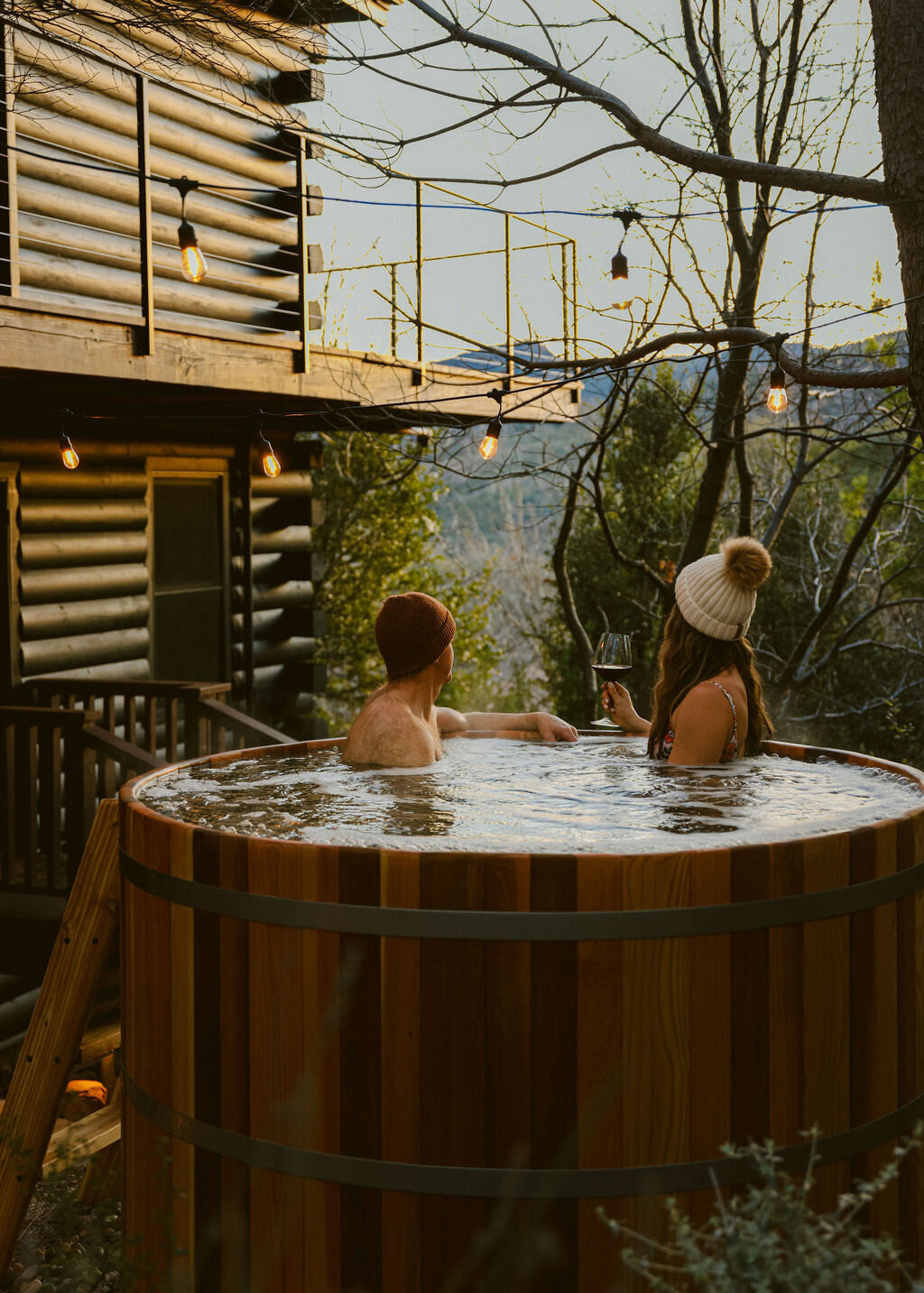 couple drinking wine in cedar hot tub with string lights in front of a-frame cabin in the mountains. Luxury boutique hotel photographer in San Diego, California