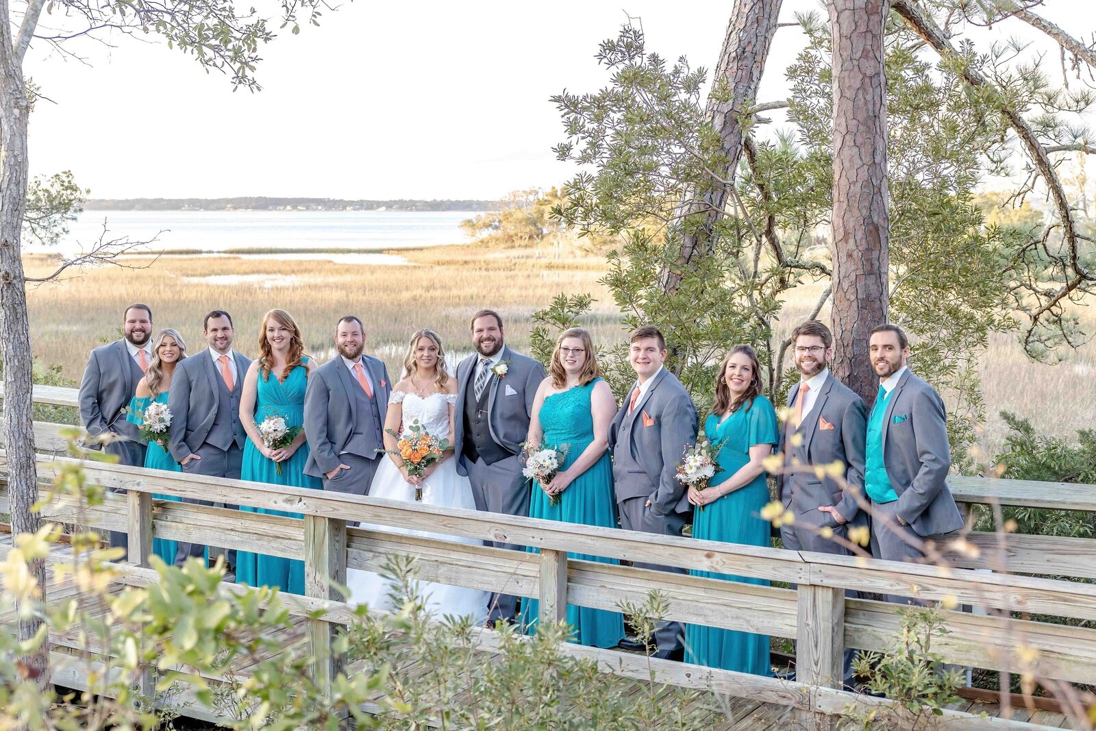The bride and groom pose with their wedding party in teal dresses and gray suits on a wooden boardwalk overlooking coastal marshes, captured during a Pine Knoll Shores Aquarium, NC wedding, blending rustic charm with waterfront beauty.