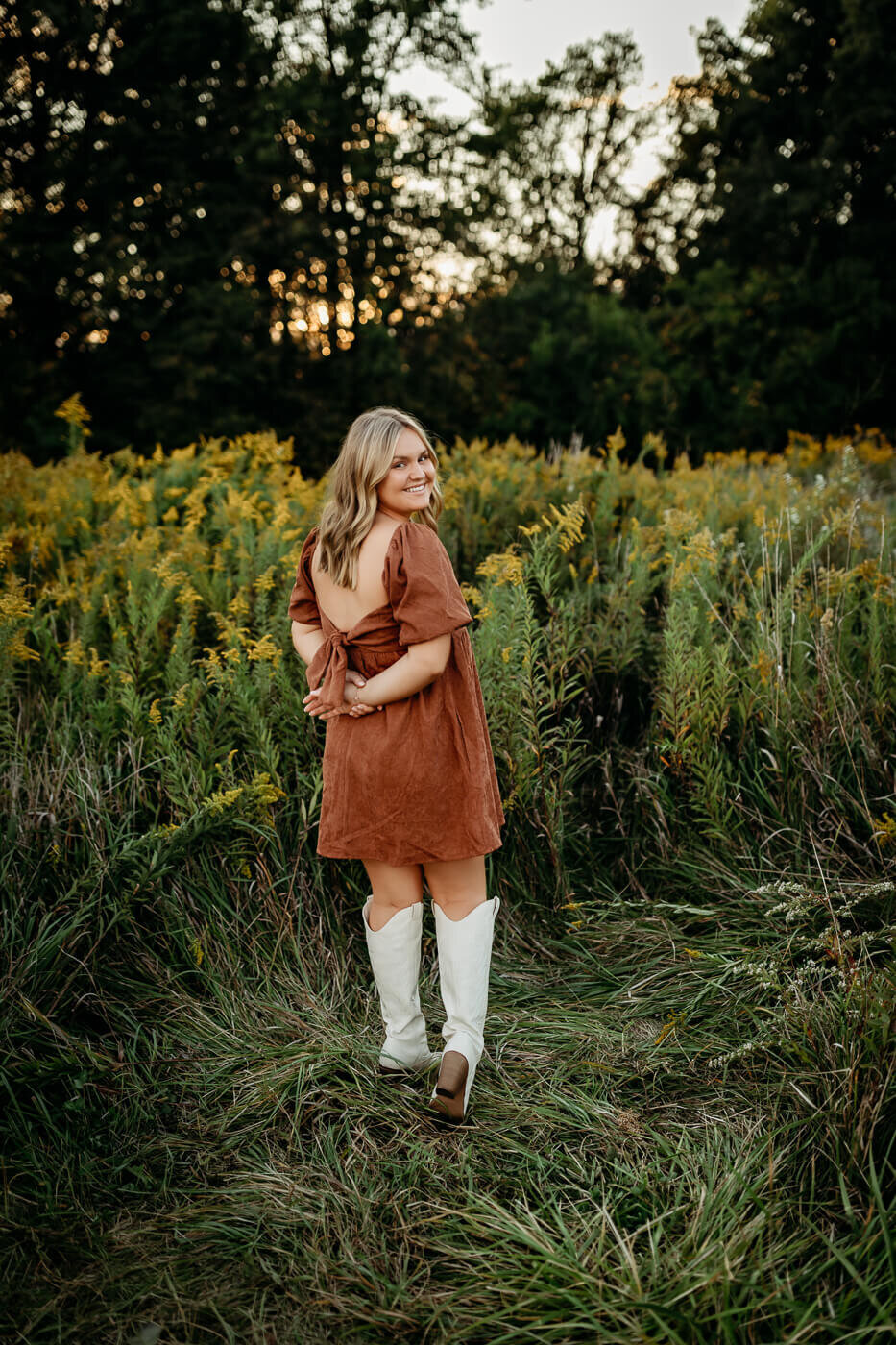 A person in a brown dress and white boots stands in a grassy field surrounded by yellow wildflowers, facing away from the camera. Tall trees are visible in the background under a dusky sky.