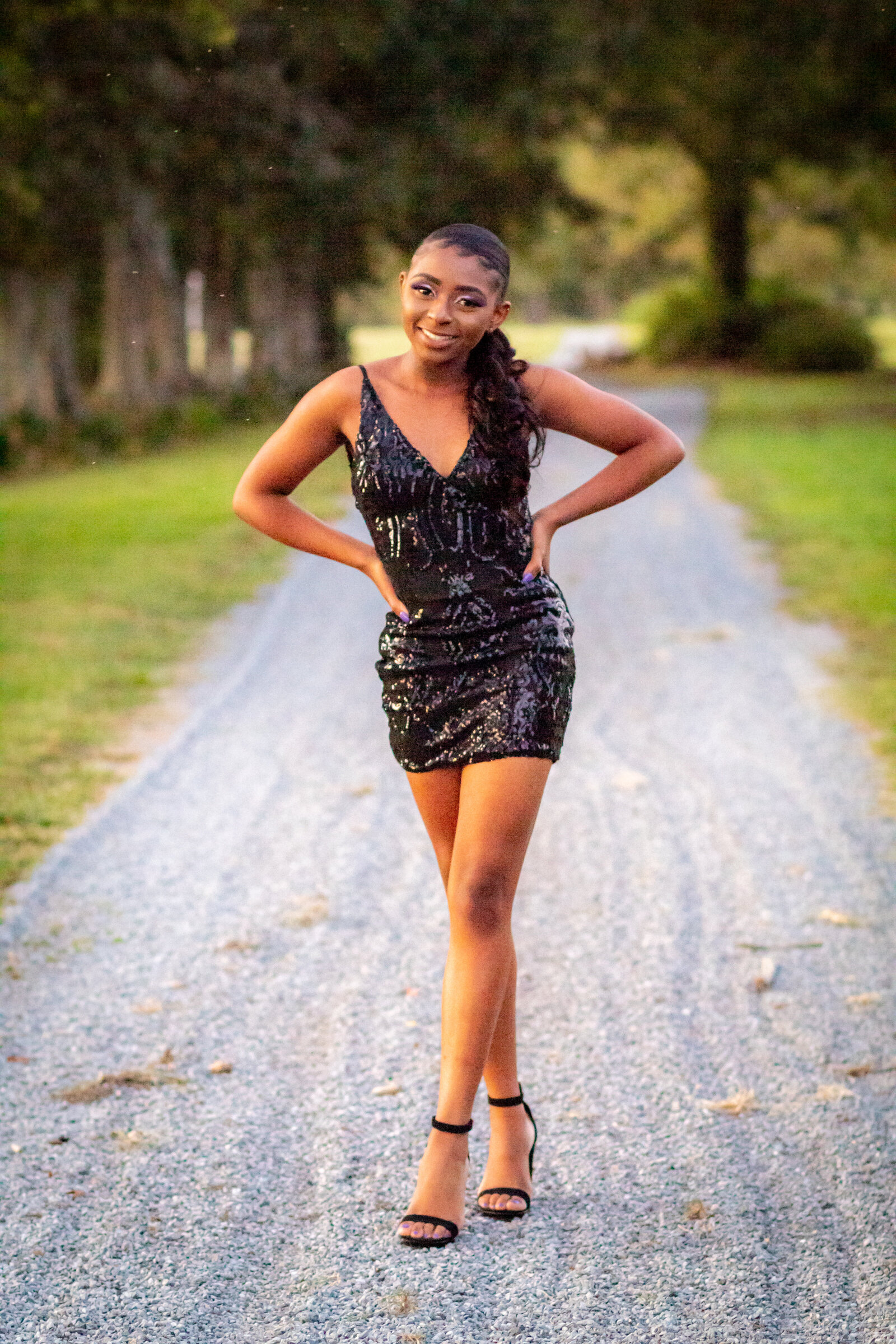 girl in black dress standing on gravel road with legs crossed with hands on hips