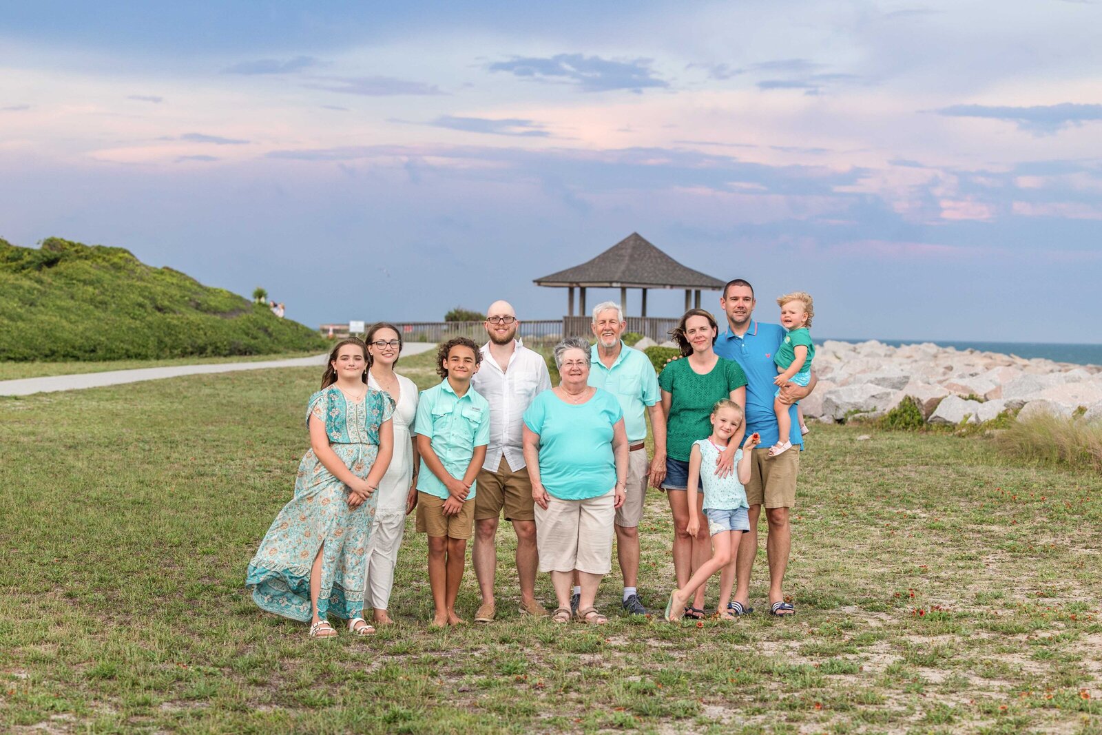 A family gathers near a gazebo and rocky coastline, dressed in teal and beige outfits. The ocean and evening sky provide a picturesque backdrop. Perfect for destination family photographer NC, showcasing scenic and joyful family reunions.
