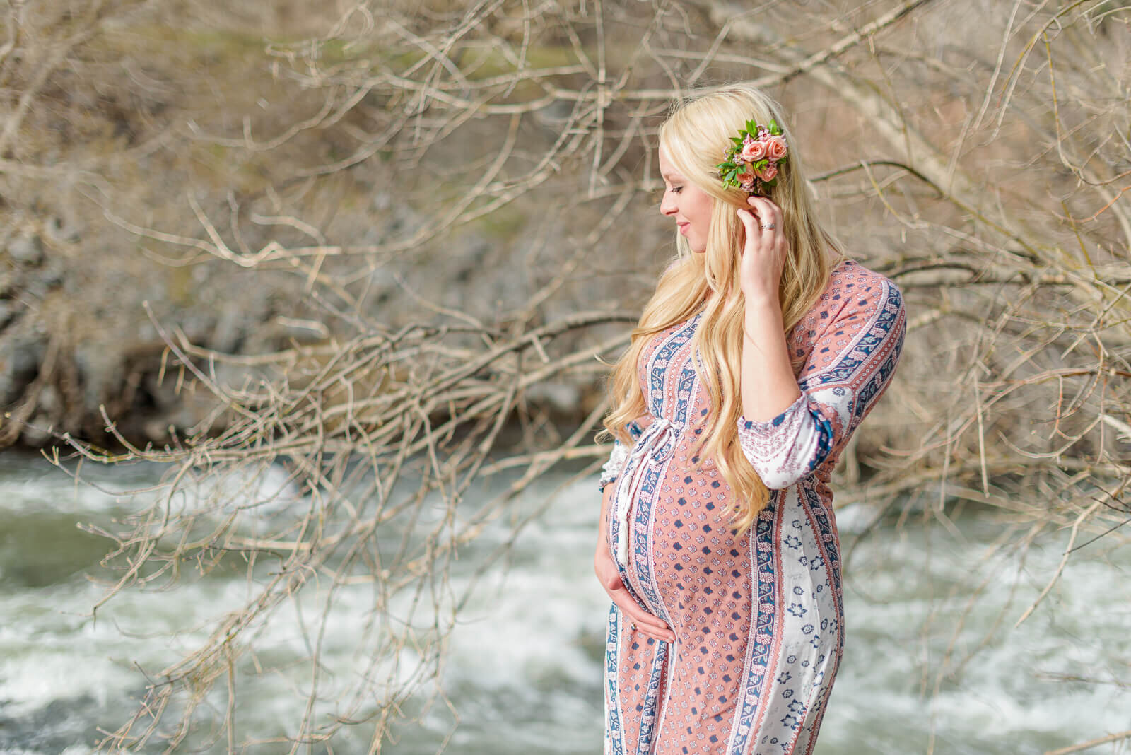 A pregnant woman wearing a pink maternity dress stands by a river in Provo Canyon. Captured by Utah maternity photographer Melissa Woodruff Photography