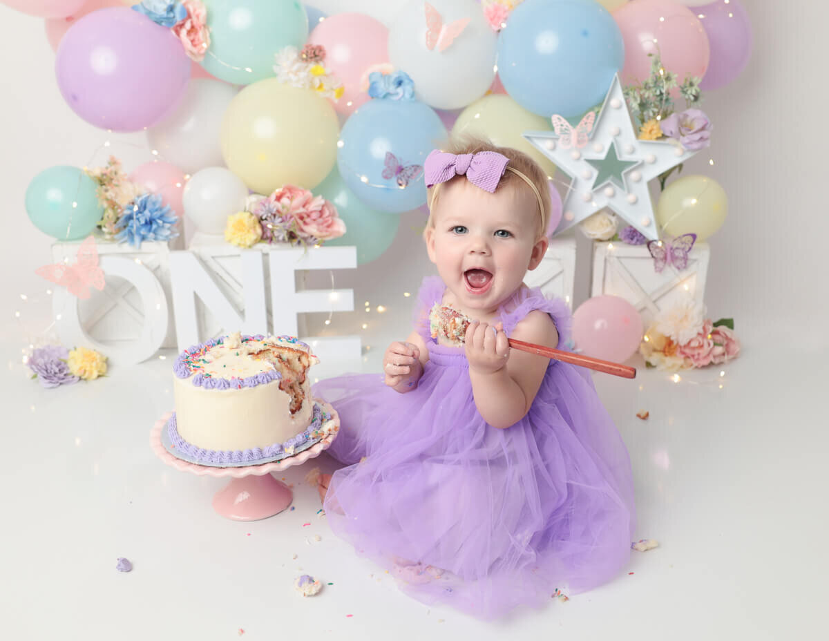 A joyful girl surrounded by vibrant pastel rainbow balloons during a studio cake smash photography session in Rochester, NY.