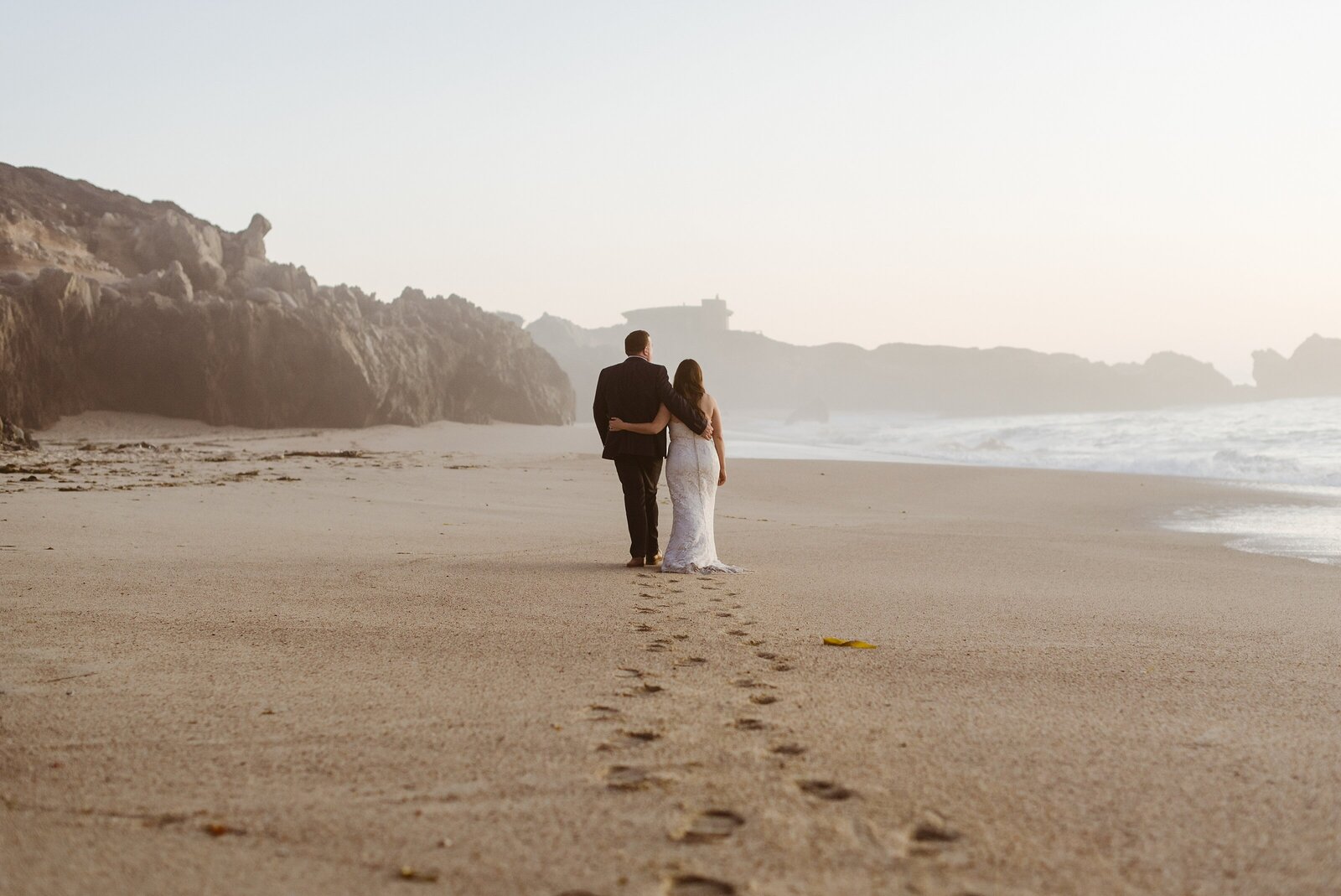Adventurous couple walks down a foggy Big Sur sandy beach with their arms around each other.