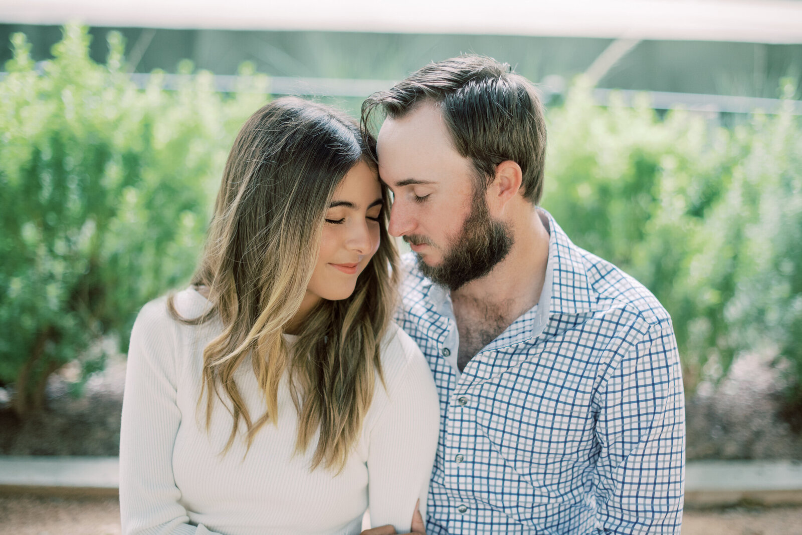 Portrait of a man and woman in a cream top and plaid dress shirt leaning into each other in front of a garden of bushes.