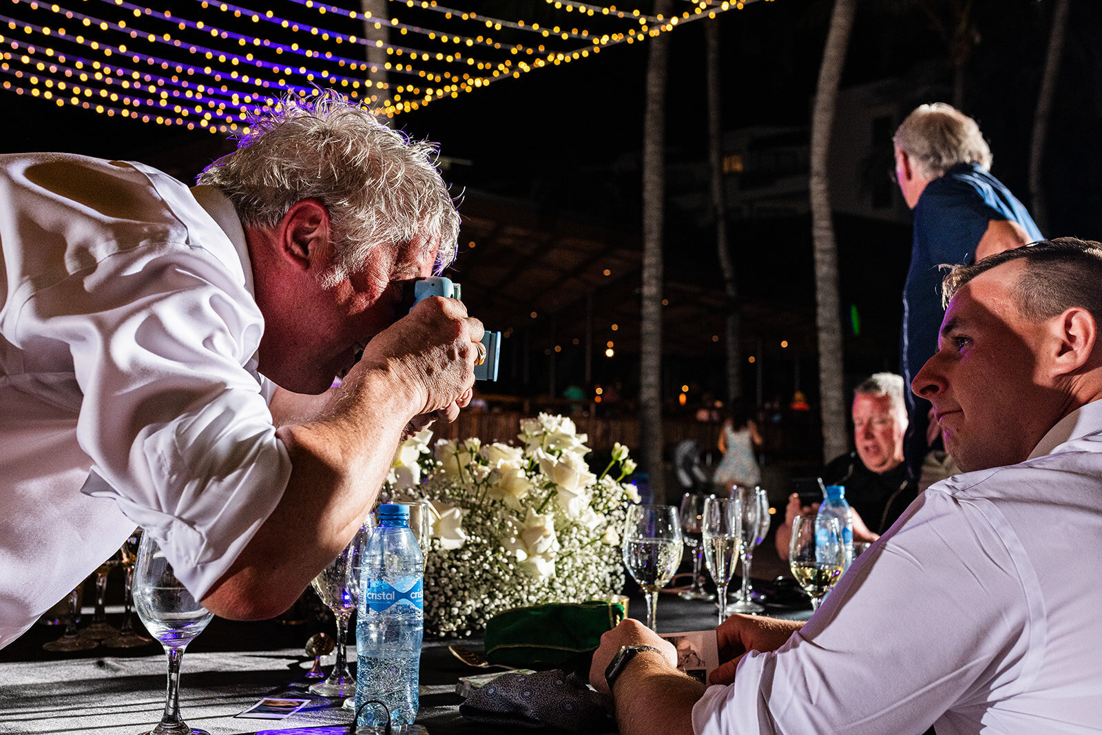 A candid moment of a guest taking a picture of the wedding details during an outdoor reception under string lights, captured by CMH Studios in a relaxed and fun atmosphere