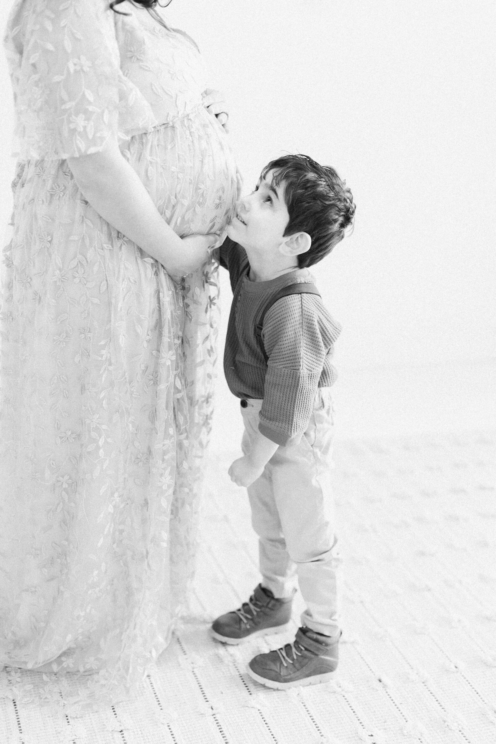 Black and white photo of little boy standing next to his pregnant mom, smiling up at her with his hand on her baby bump by NH newborn photographer Fieldstone Studio.